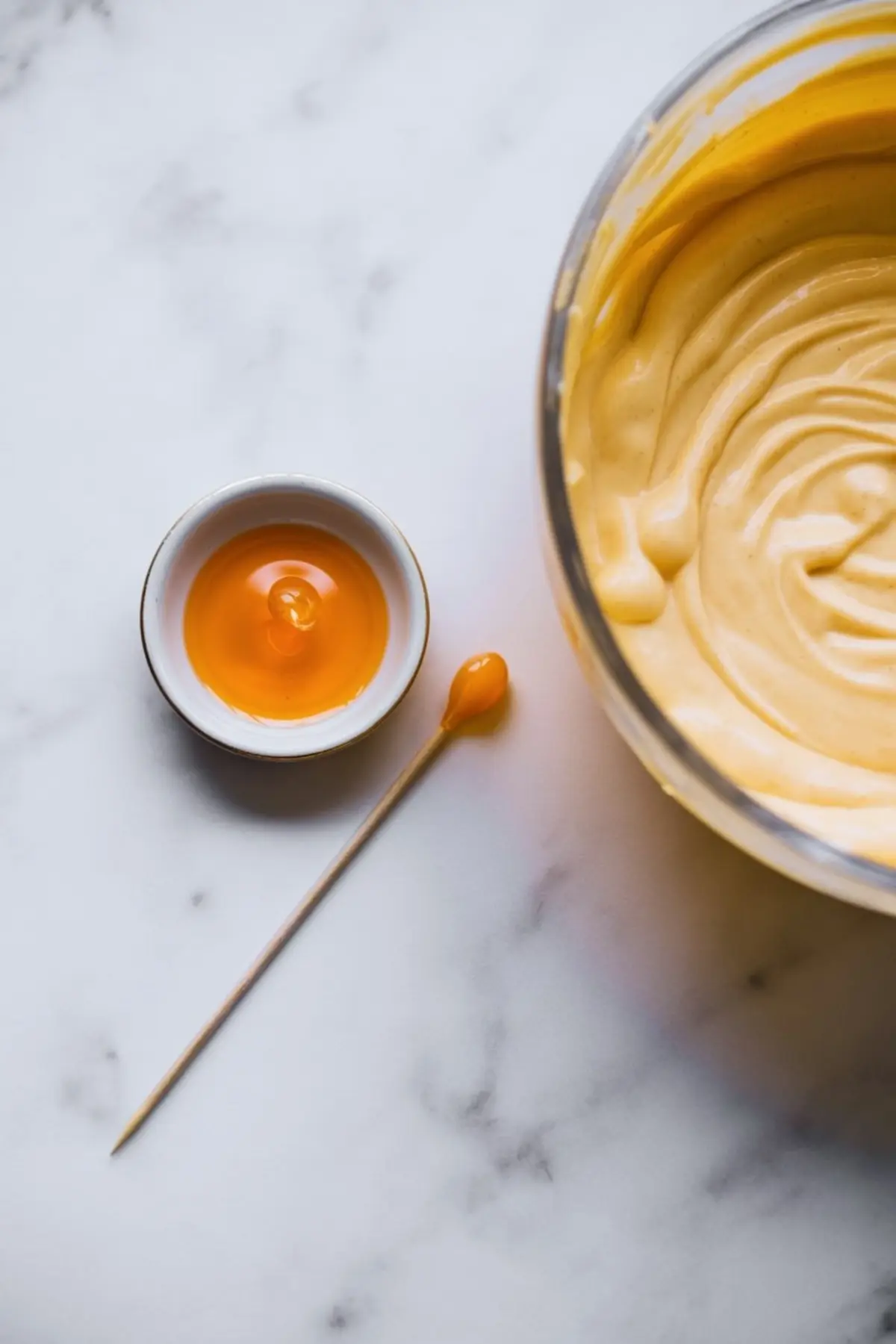 Creamy pumpkin cheesecake batter in a glass bowl with a small dish of orange food coloring and a cocktail stick on a marble countertop.