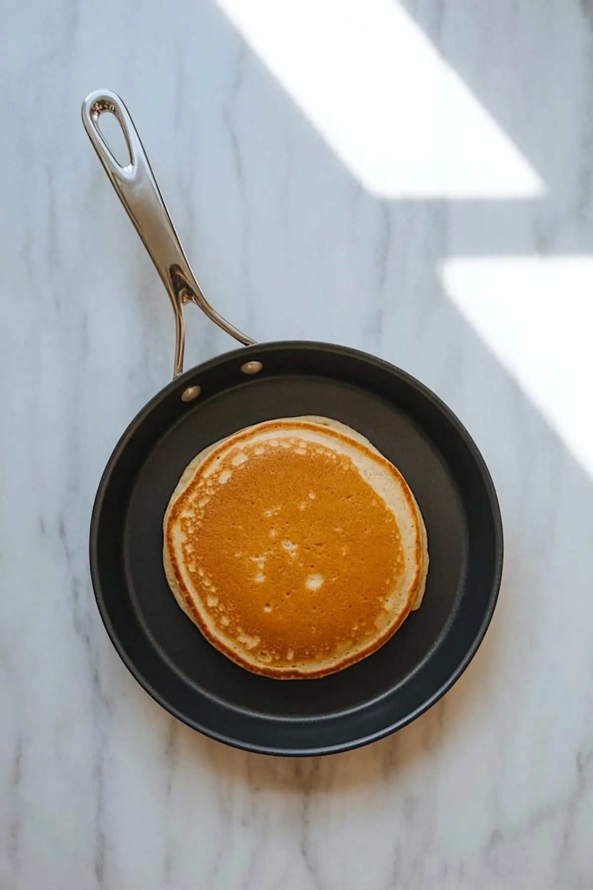Golden brown pancake cooking in a non-stick skillet on a white marble surface, with even edges and a fluffy center.