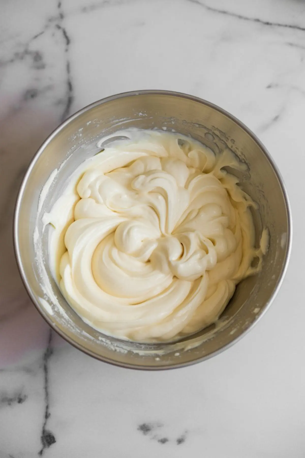 Stainless steel bowl filled with whipped cheesecake batter, swirled to a creamy and fluffy texture, resting on a white marble background.