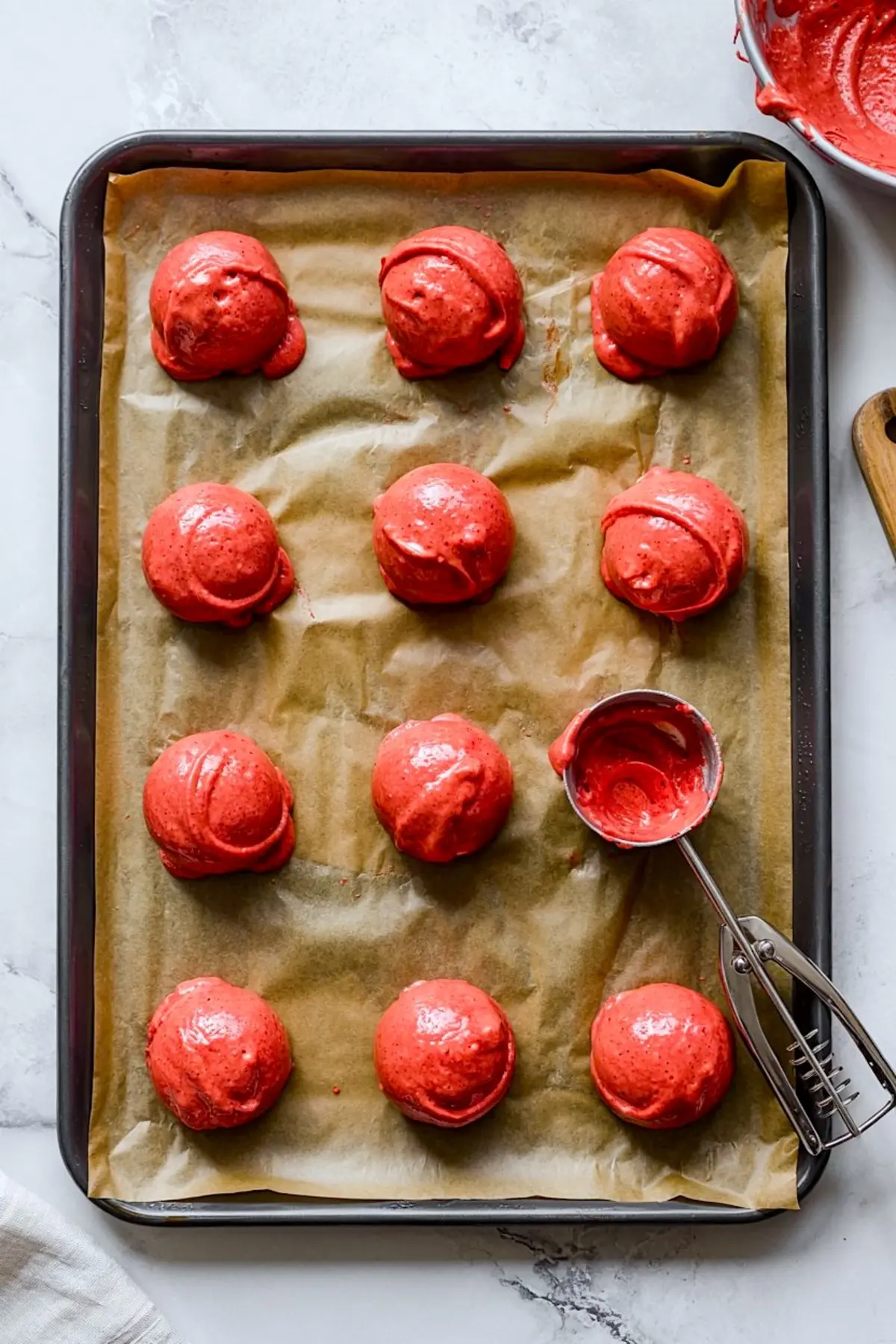 Round scoops of red velvet batter are placed on a parchment-lined baking sheet, ready for baking, with a metal cookie scoop resting beside them.