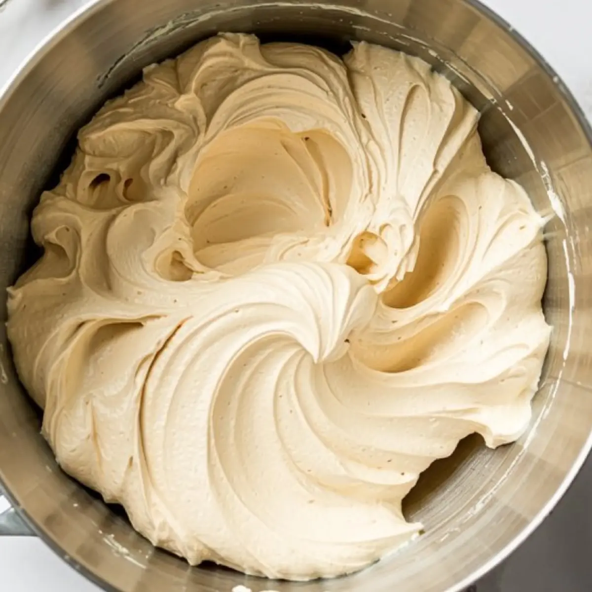 Fluffy vanilla buttercream frosting swirls in a large stainless steel mixing bowl, showing a smooth and airy texture.