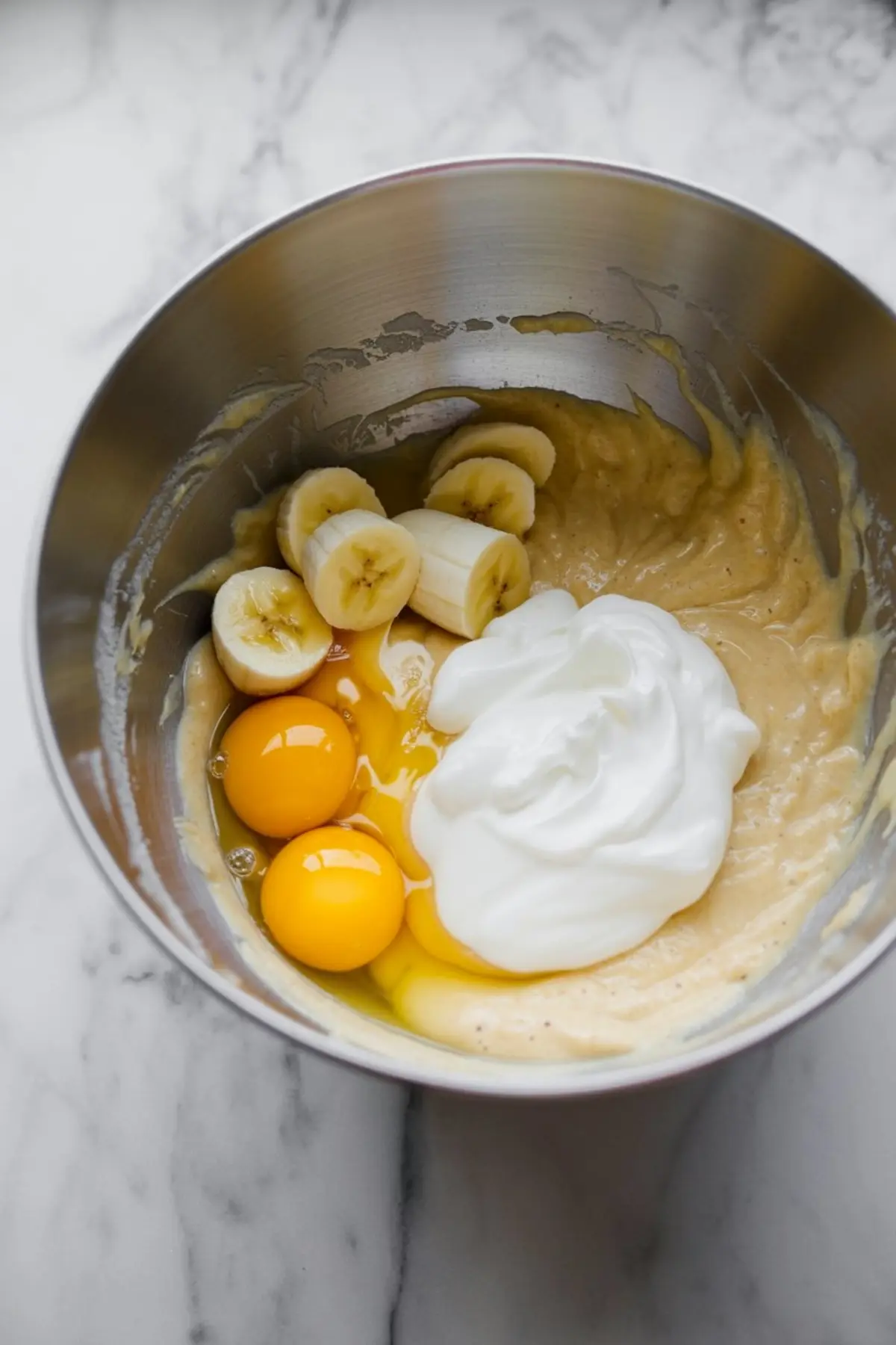 Metal mixing bowl with banana bread batter, showcasing sliced bananas, eggs, and Greek yogurt just added before mixing, on a marble countertop.