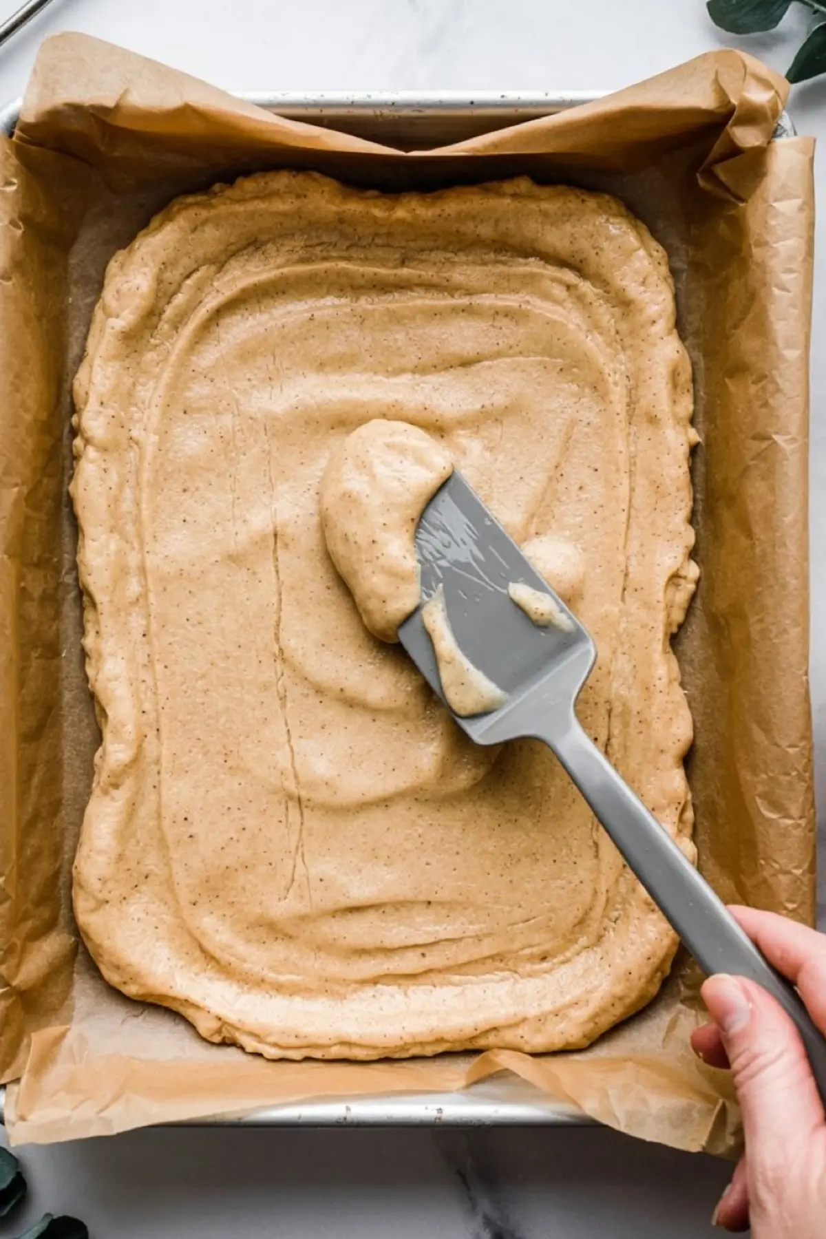 Unbaked snickerdoodle batter being evenly spread in a parchment-lined baking pan using a spatula.