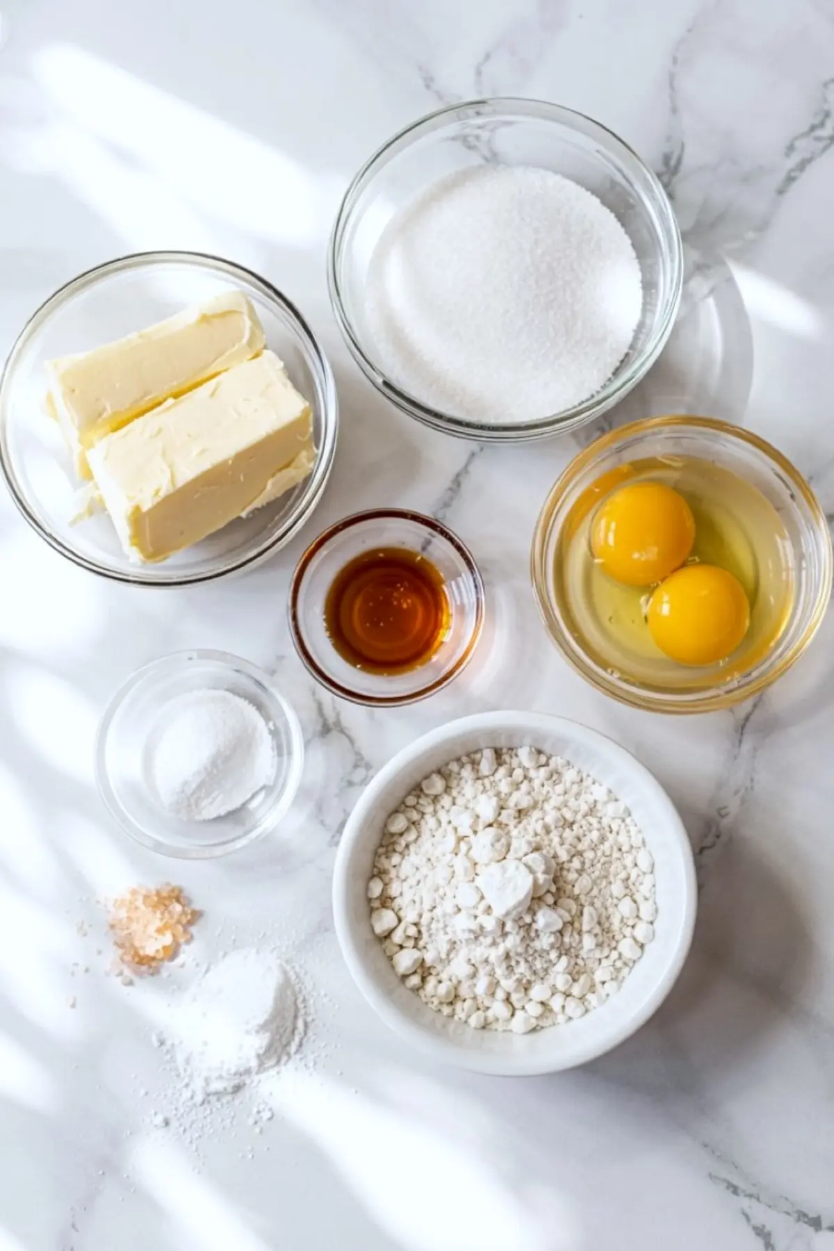 Overhead view of baking ingredients on a marble surface, including butter, sugar, eggs, vanilla extract, flour, baking soda, cream of tartar, and salt arranged in glass and ceramic bowls.