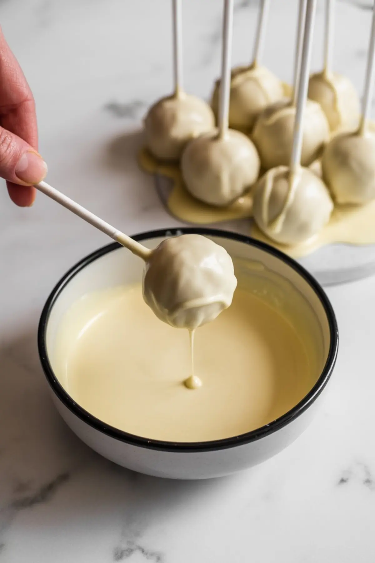 Hand dipping a round cake pop into a bowl of melted white chocolate with several already-coated cake pops standing upright in the background on parchment paper.
