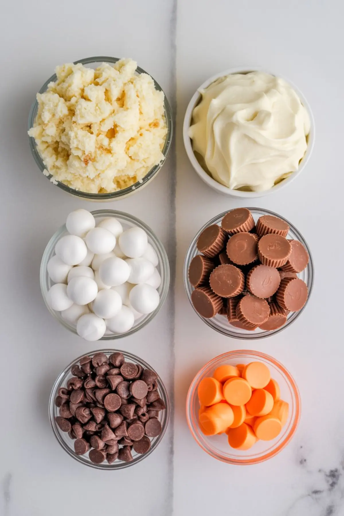 Overhead view of ingredients for snowman cake pops, including crumbled cake, frosting, white candy melts, mini peanut butter cups, chocolate chips, and orange candy melts arranged in small bowls on a white surface.
