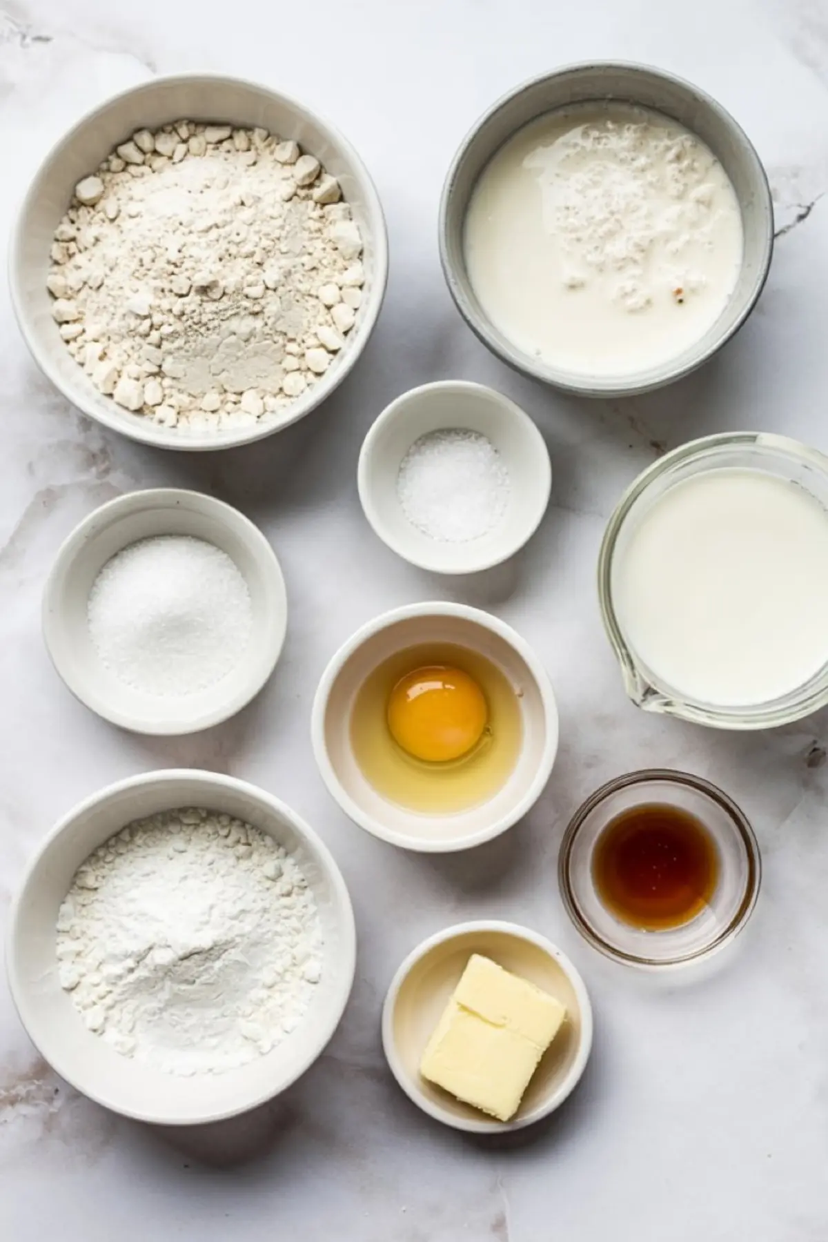 Flat lay of pancake ingredients including flour, sugar, baking powder, milk, buttermilk, egg, butter, vanilla extract, and salt arranged in small bowls on a marble surface.