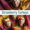 Strawberries coated in chocolate and styled as turkey faces using candy eyes and fondant beaks, placed in paper cups on a plaid fall napkin for a festive Thanksgiving treat display.