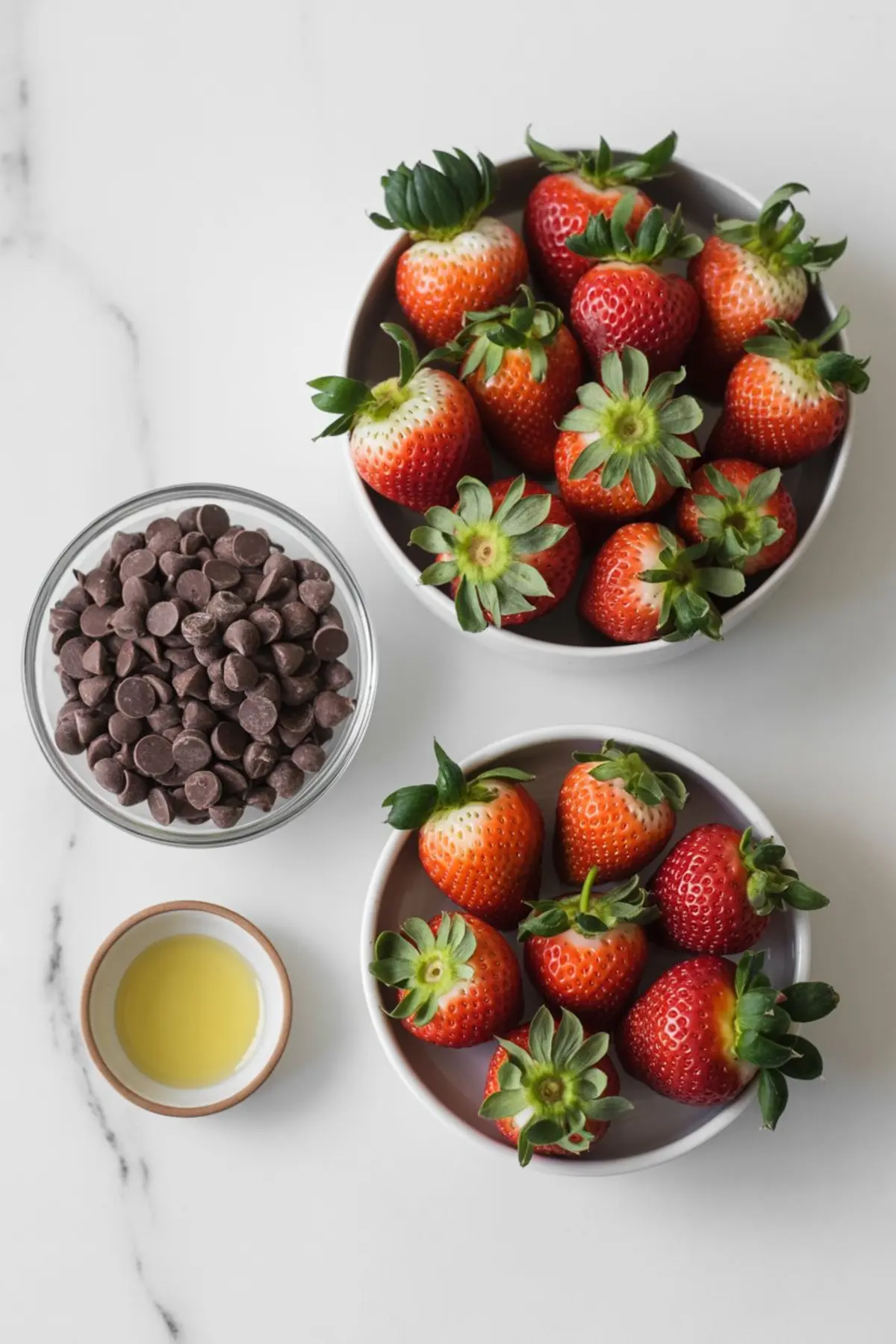 Flat lay of Thanksgiving treat ingredients showing fresh strawberries in bowls, a dish of chocolate chips, and a small bowl of oil on a white surface.