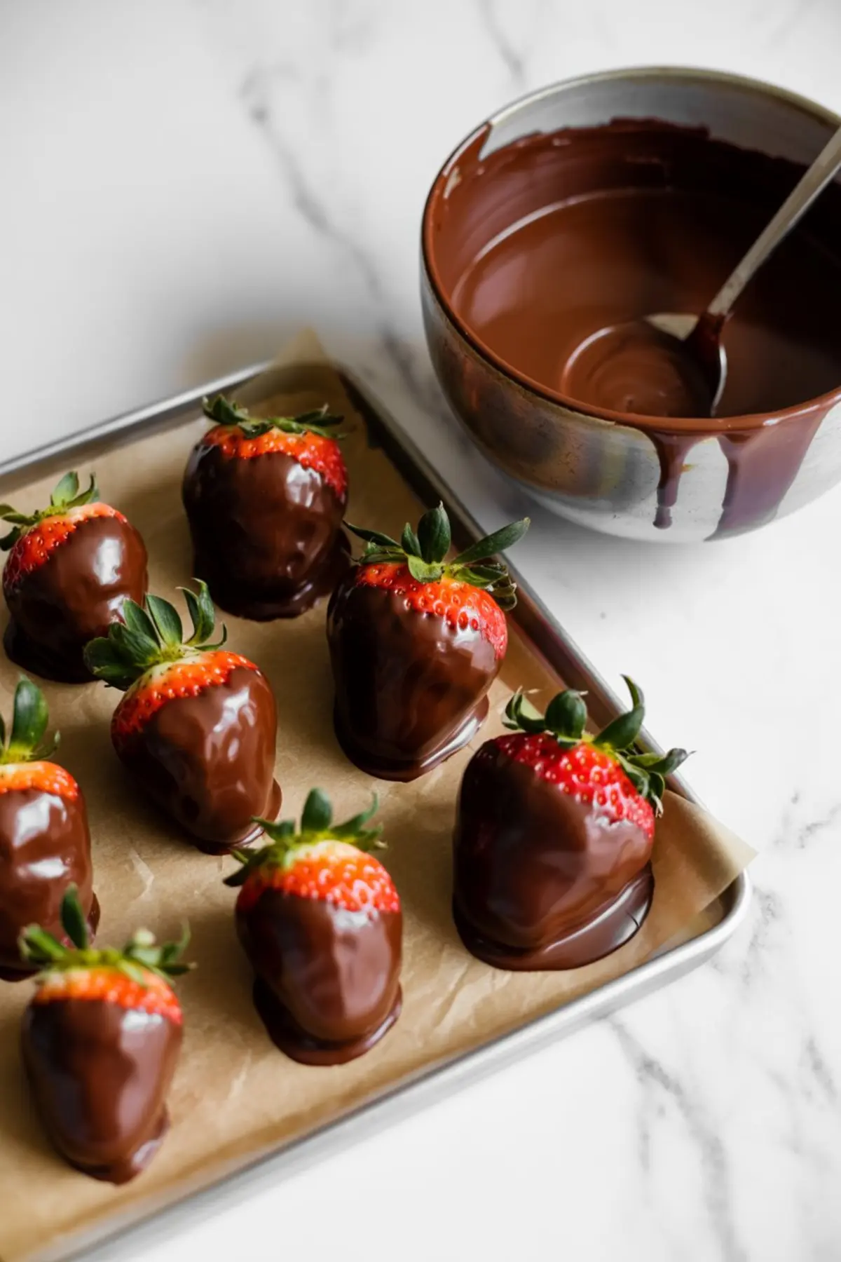 Tray of strawberries partially dipped in melted chocolate, set on parchment paper next to a mixing bowl with a spoon, cooling and setting for holiday treat decorating.