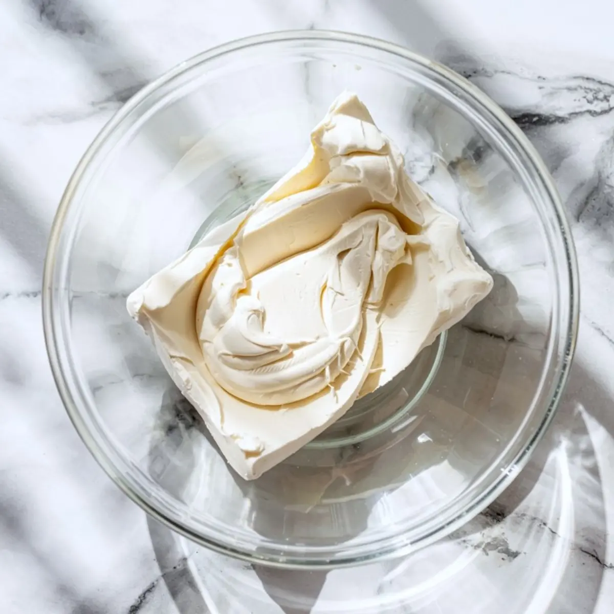 Block of softened cream cheese in a glass mixing bowl on a white marble surface, ready for blending dessert dip recipes.

