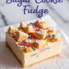 Square piece of sugar cookie fudge topped with colorful sprinkles and cookie chunks, served on white parchment paper with a smooth, creamy texture.