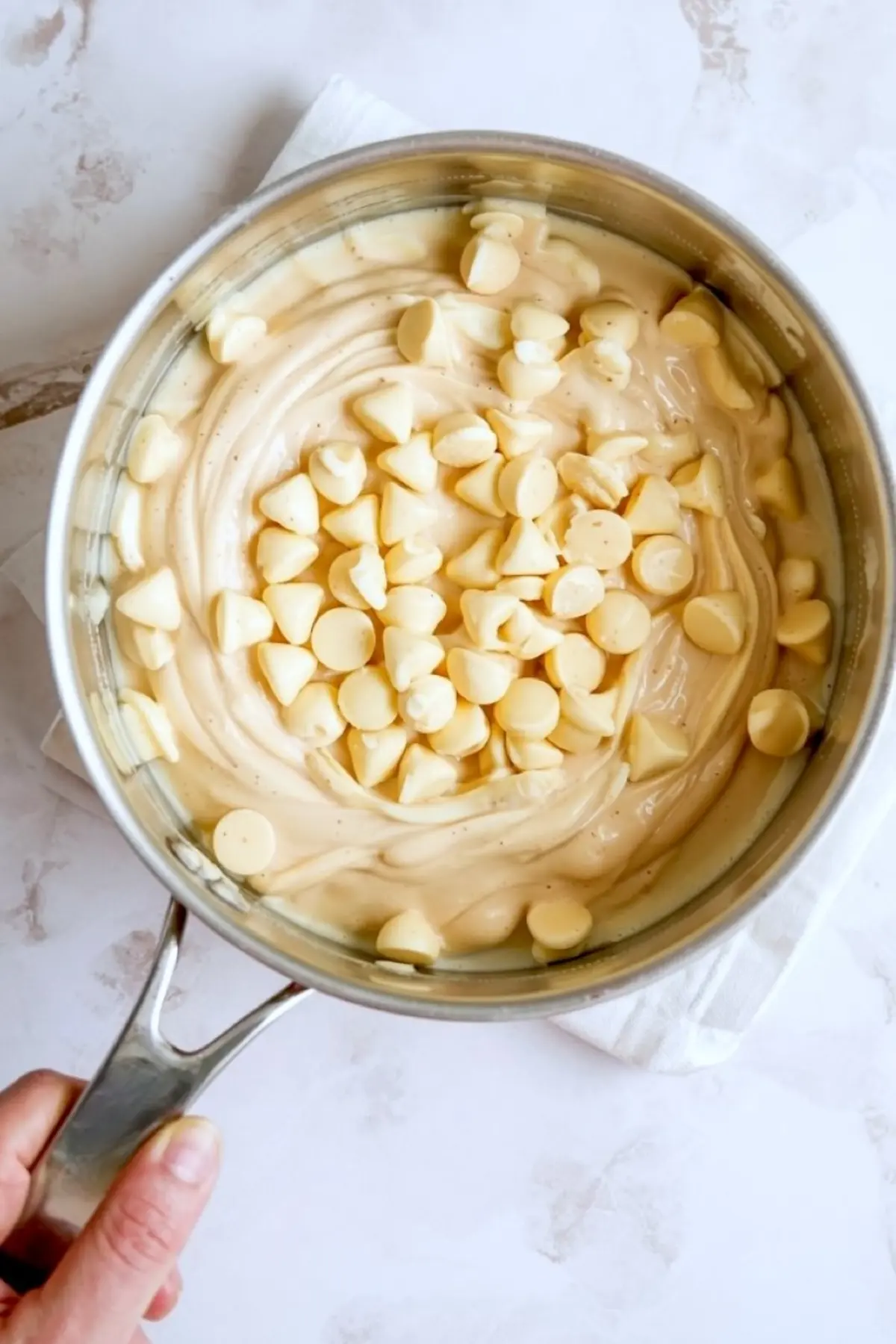 Melted white chocolate chips folded into creamy vanilla fudge mixture in a stainless steel saucepan, shown from above on a white background.