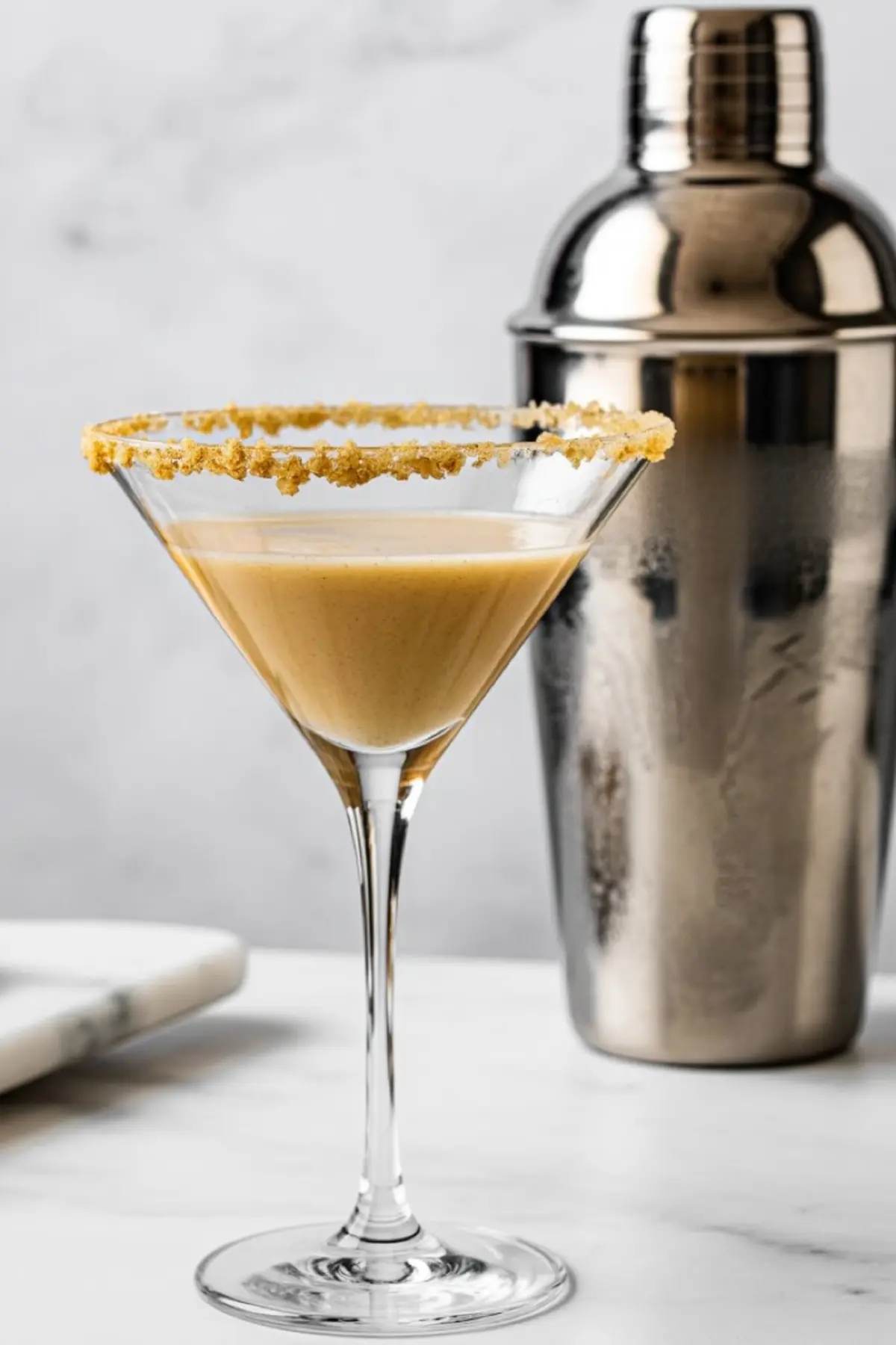 A classic martini glass filled with a creamy tan-colored sugar cookie martini, garnished with a crushed cookie rim. A stainless steel cocktail shaker stands in the background on a marble surface.
