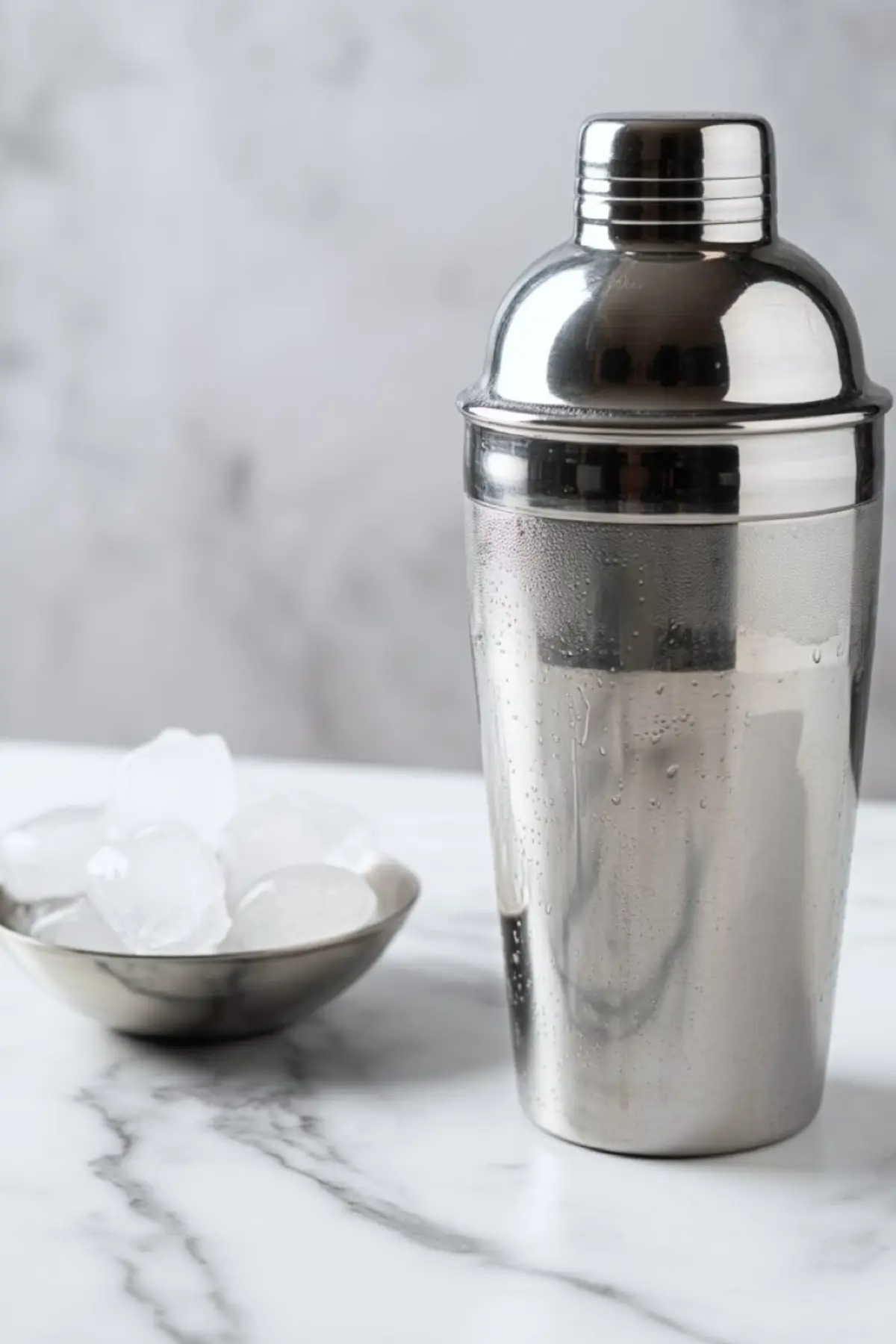 A condensation-covered stainless steel cocktail shaker beside a small metal bowl filled with ice cubes on a white marble countertop.