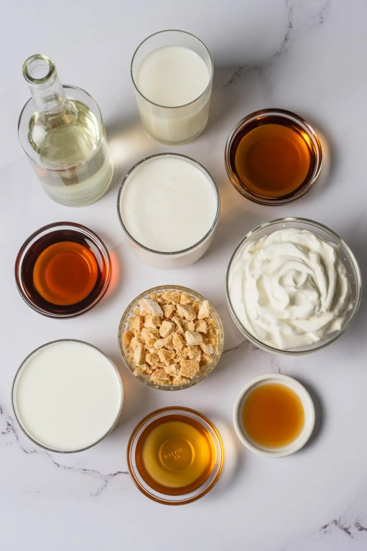Flat lay of sugar cookie martini ingredients on a white surface, including vodka, milk, cream, whipped topping, vanilla extract, and crushed cookies in glass bowls and cups.