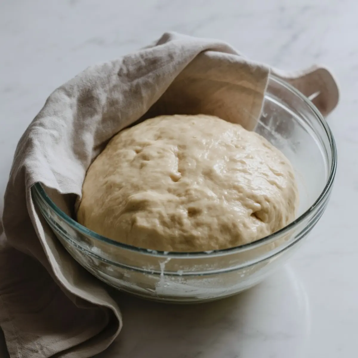 Risen focaccia dough in a glass bowl partially covered with a beige cloth, ready for shaping or transferring to the baking pan.