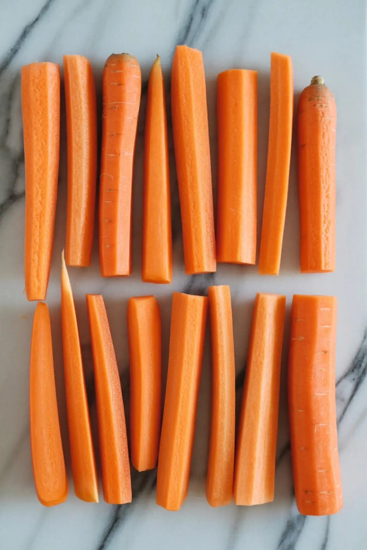 Close-up of fresh carrots sliced into long sticks, neatly aligned on a white and gray marble surface.