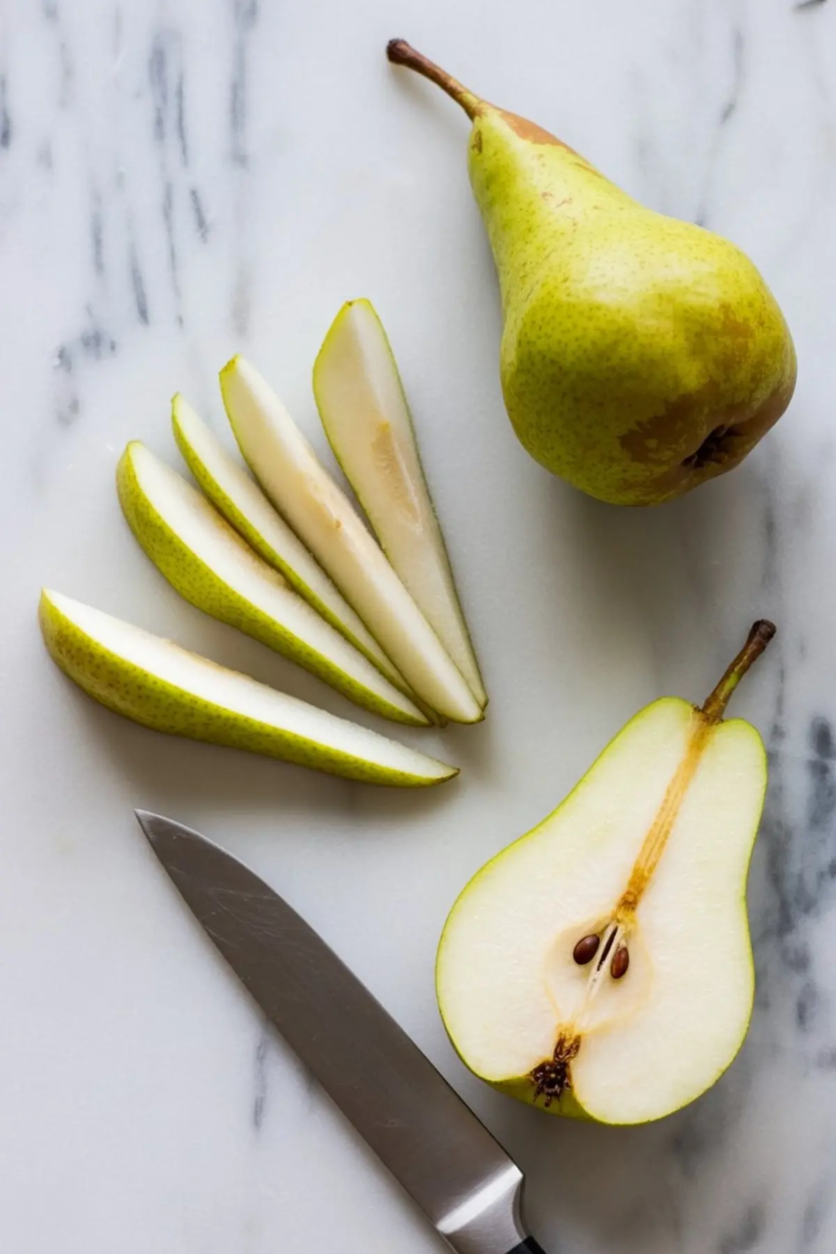 Green pear sliced into thin wedges on a marble surface, displayed with a knife and a halved pear showing the seeds.