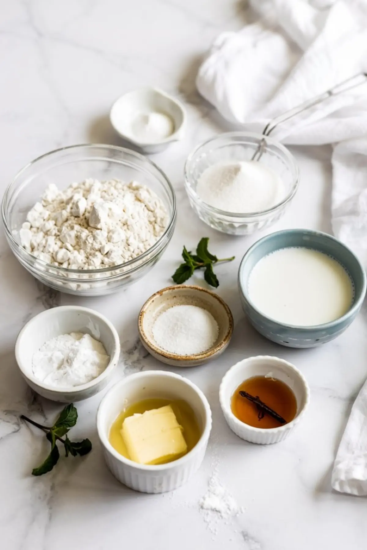 Flat lay of ingredients for vanilla mug cake including flour, sugar, baking powder, salt, butter, vanilla extract, and milk, arranged in bowls on a white marble surface.
