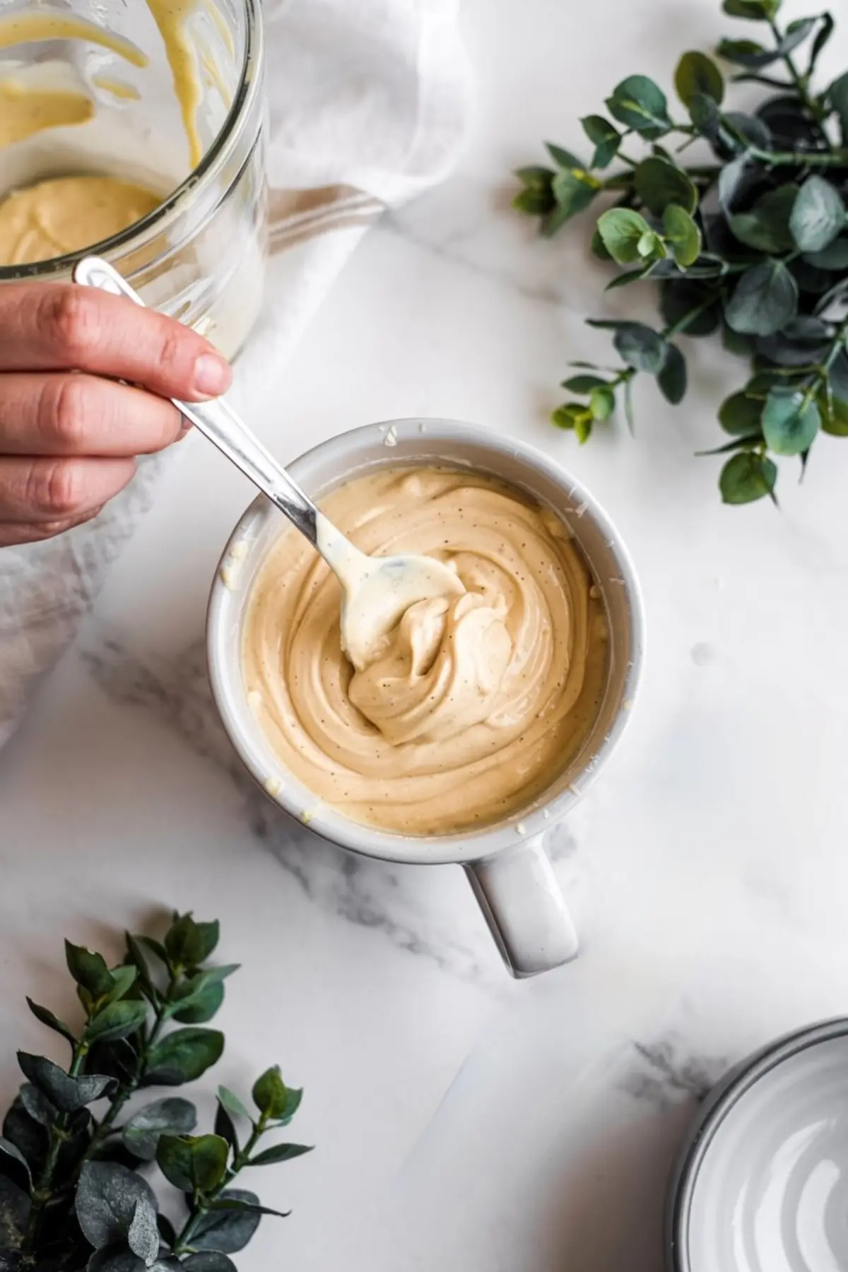 Hand stirring vanilla mug cake batter inside a white mug, with creamy, smooth texture ready for microwaving, surrounded by green leafy décor.
