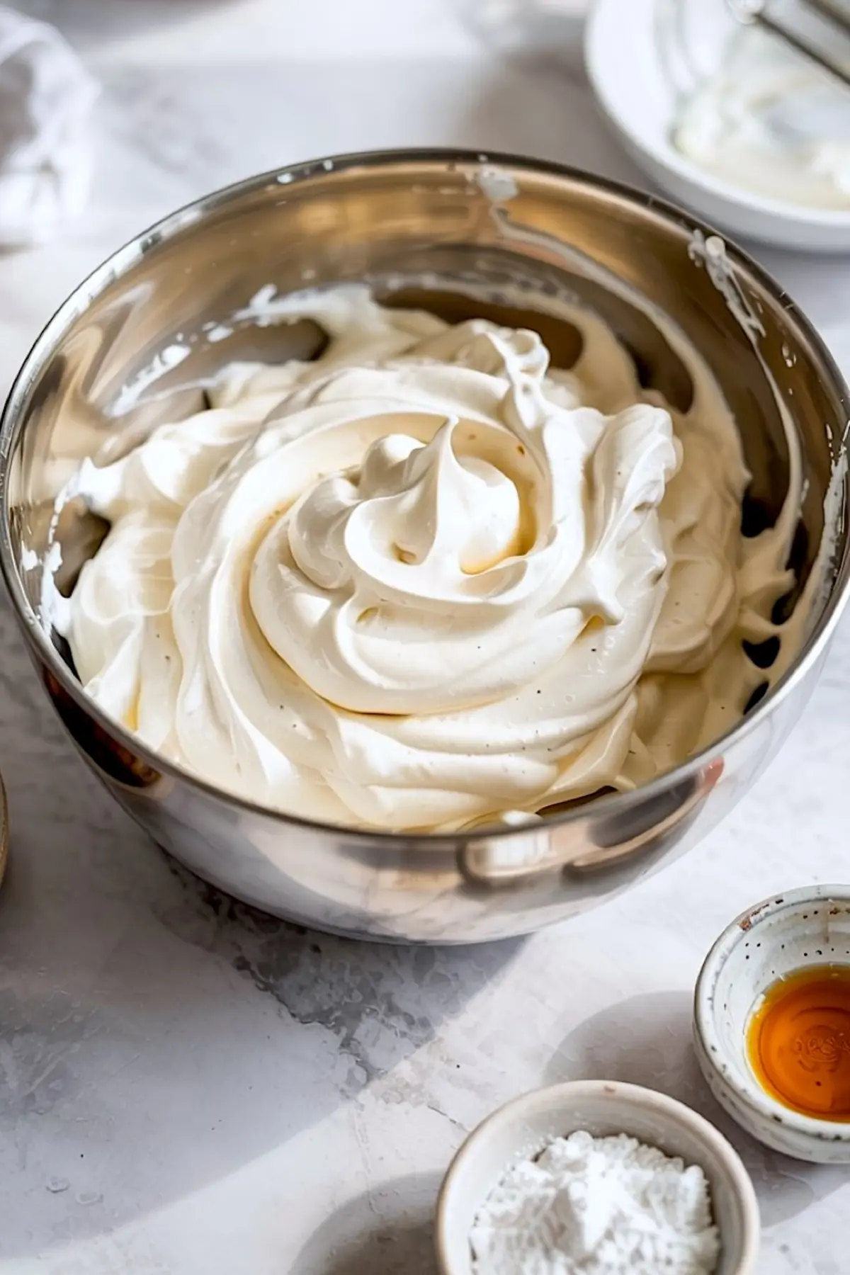 Glossy metal mixing bowl filled with whipped vanilla frosting, surrounded by small ceramic bowls containing vanilla extract and powdered sugar.
