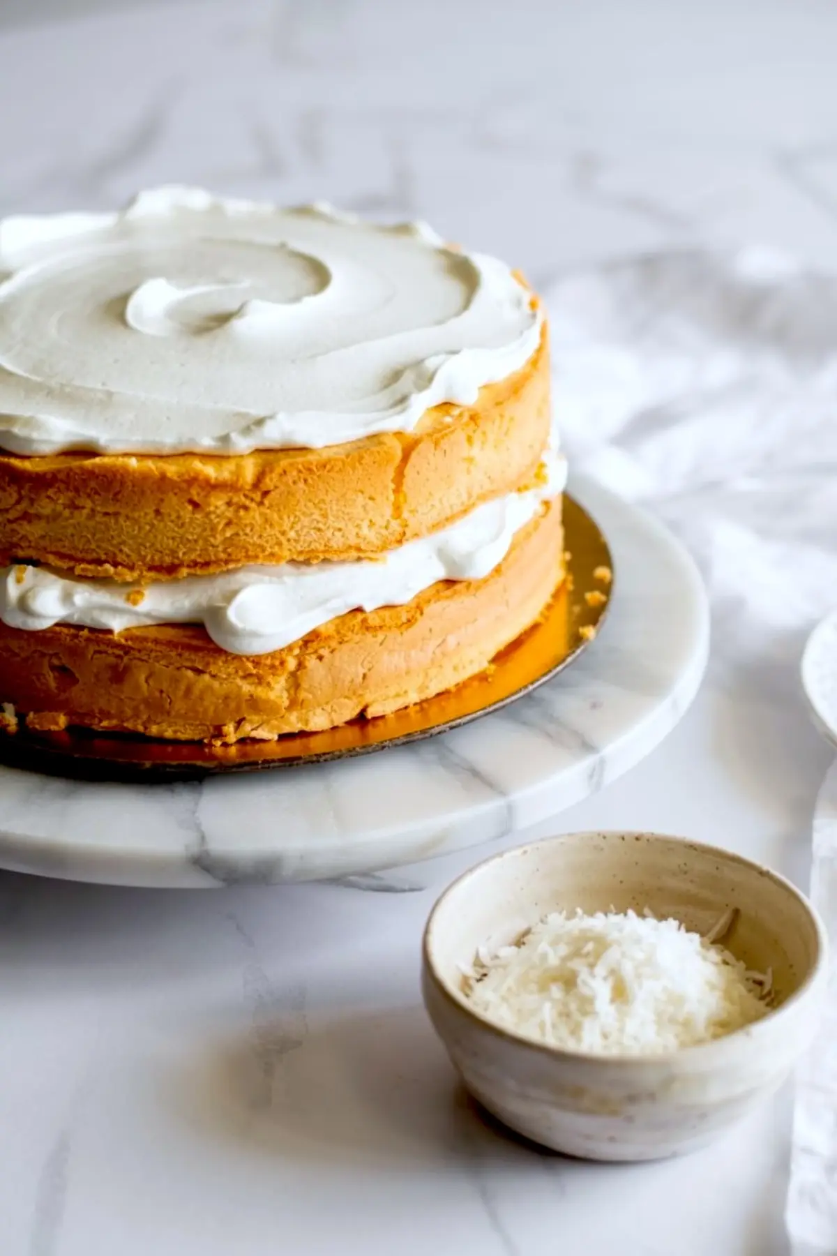 Unfinished vanilla sponge cake with two thick layers and a swirl of whipped frosting on top, set on a marble cake stand with a small bowl of shredded coconut beside it.
