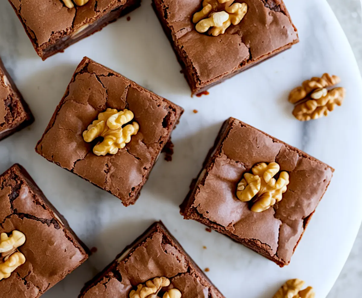Fudgy walnut brownies with crackly tops, each topped with a whole walnut, displayed on a marble platter.
