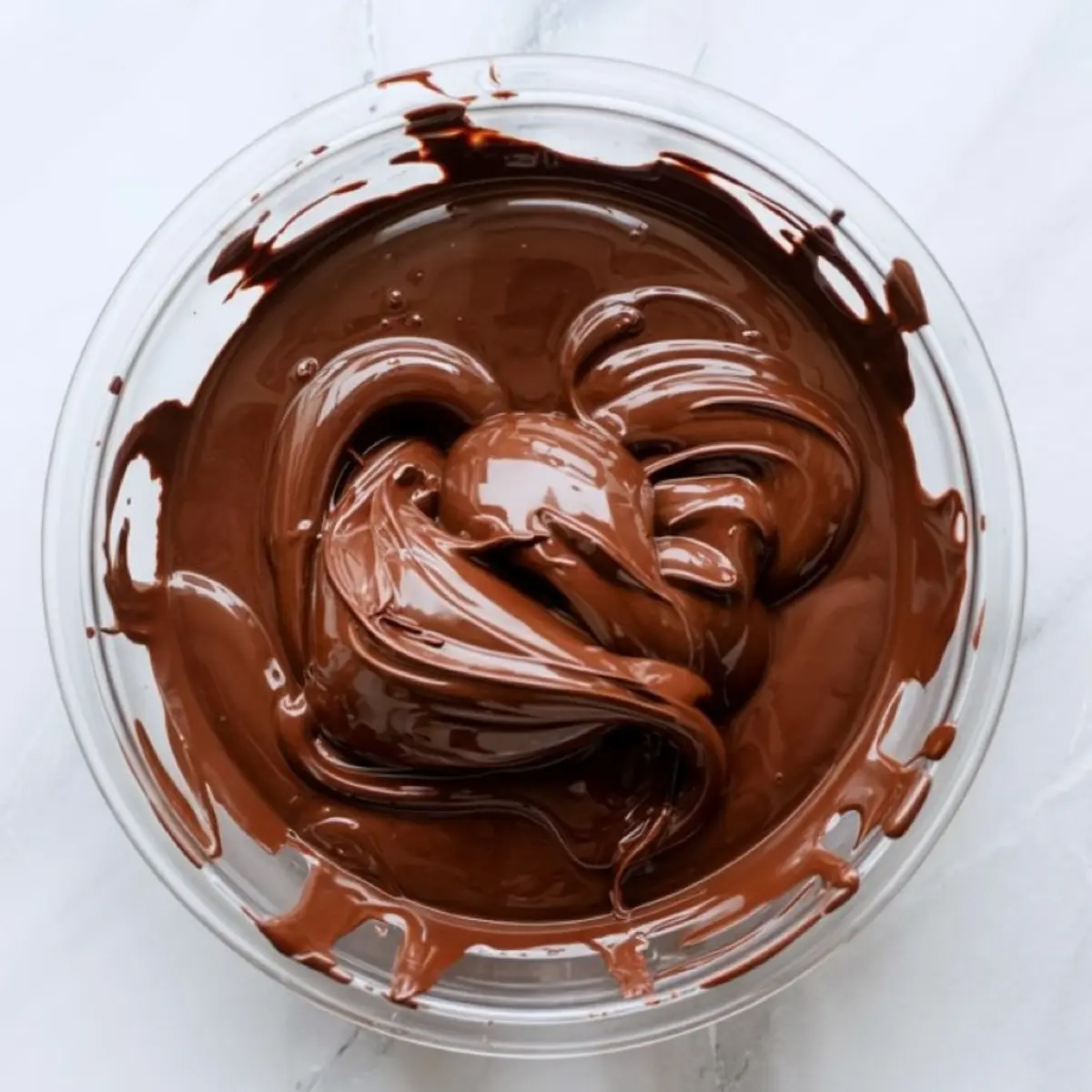 Glossy melted chocolate swirled in a clear glass bowl on a white marble surface.
