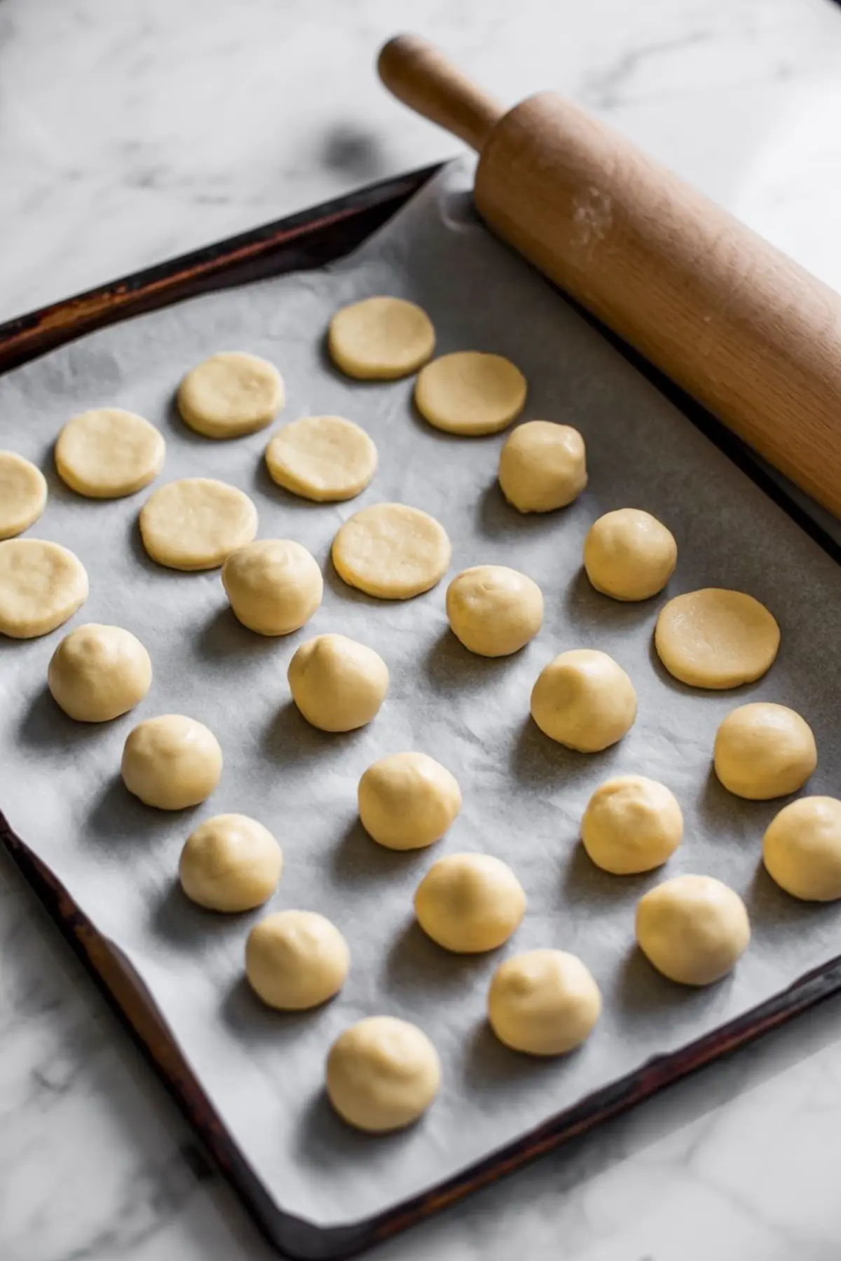 Unbaked walnut cookie dough rolled into balls and pressed into discs on a parchment-lined baking tray with a wooden rolling pin beside it.
