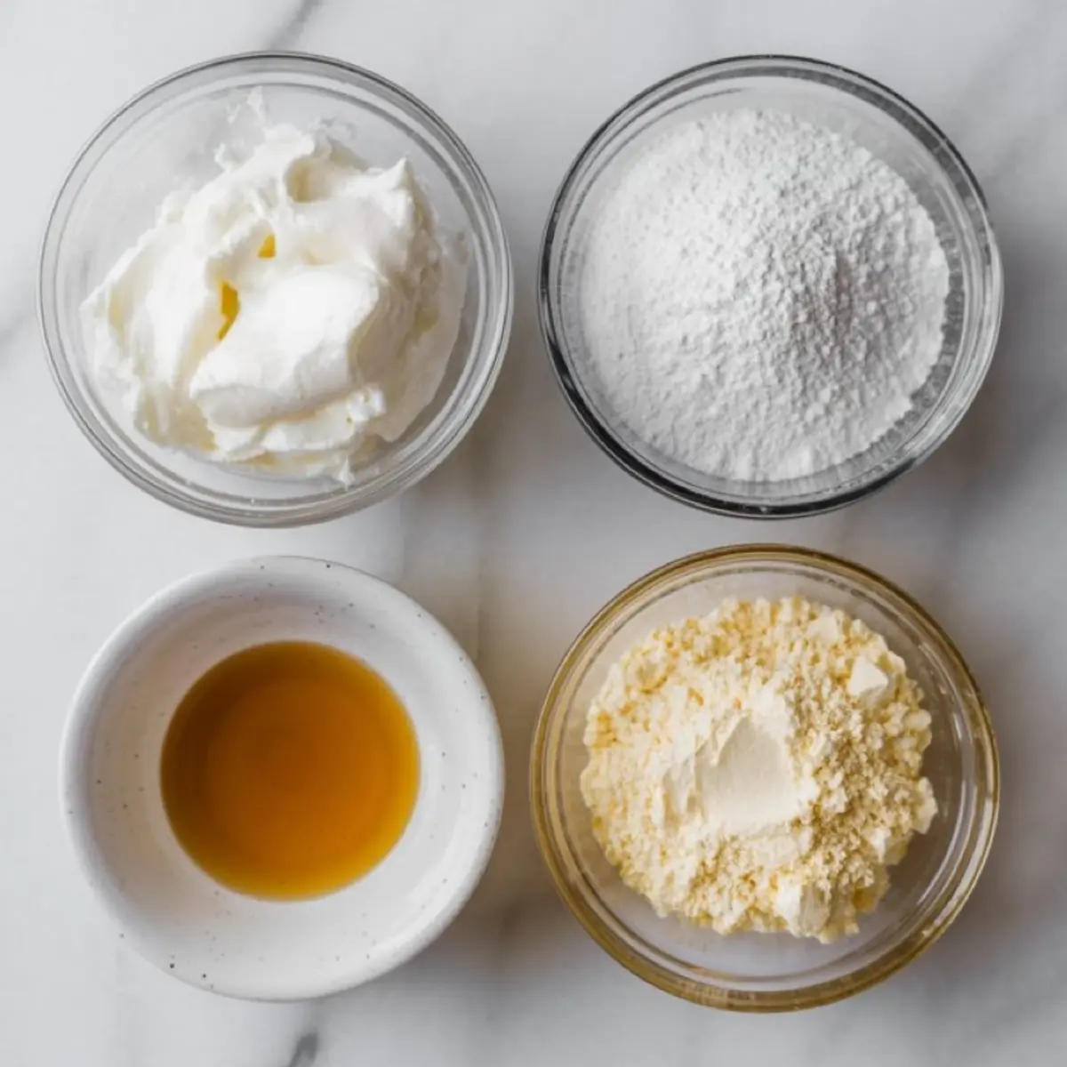 Top-down view of whipped cream frosting ingredients in separate bowls, including powdered sugar, vanilla extract, whipped topping, and instant pudding mix on a marble surface.
