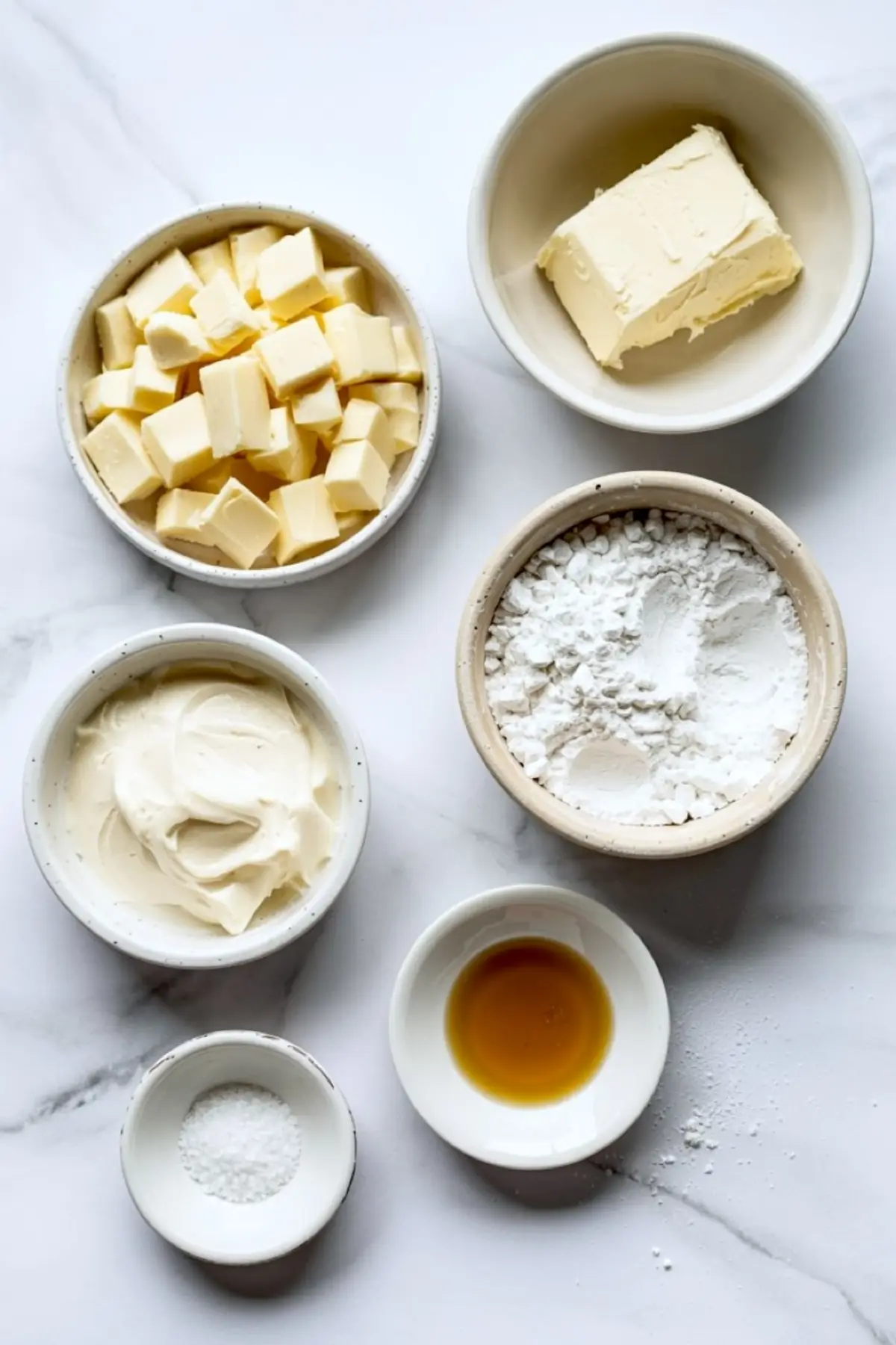 Flat lay of white chocolate frosting ingredients on a white marble surface, including cubed butter, cream cheese, powdered sugar, vanilla extract, salt, and a block of softened butter.
