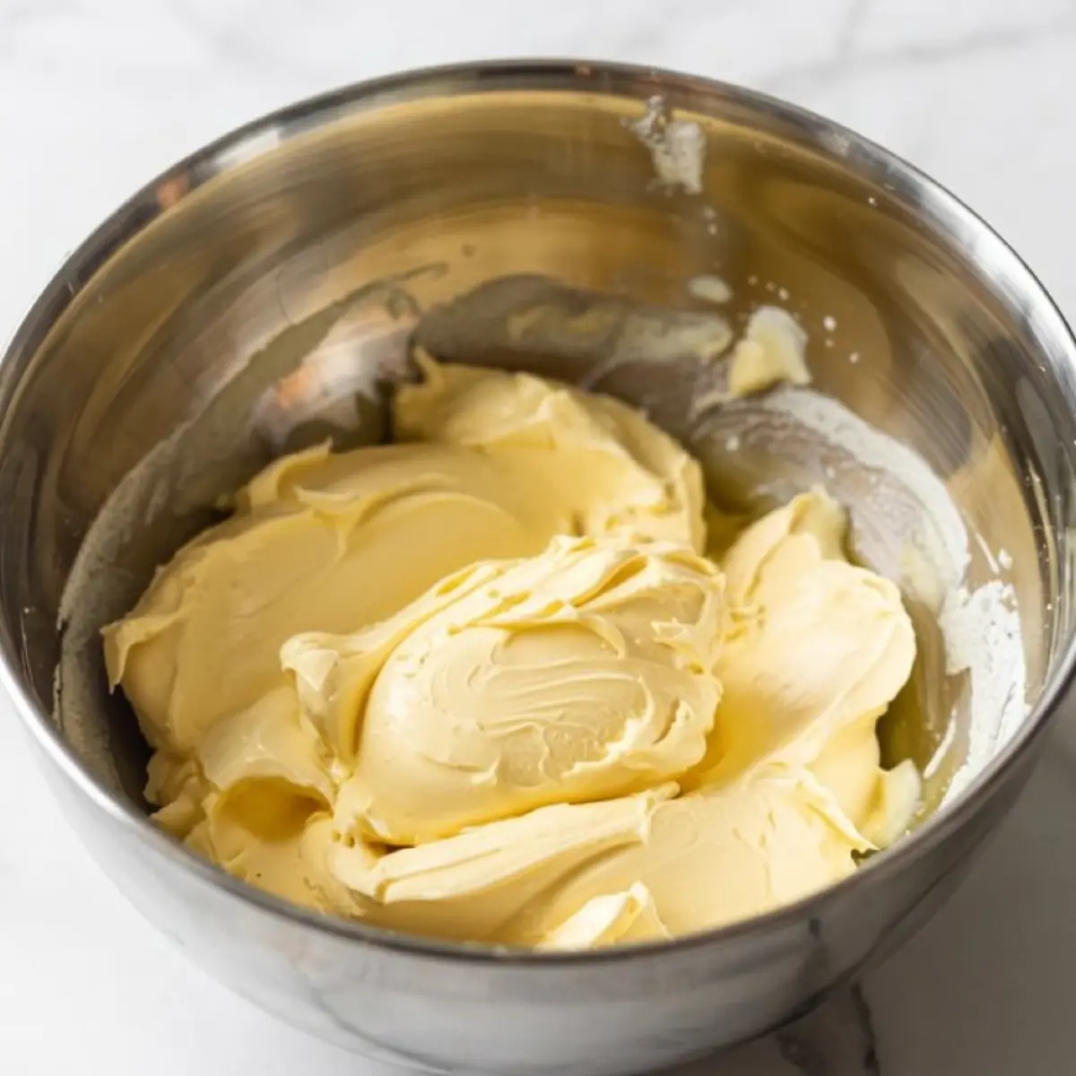 Creamy white chocolate frosting whipped to a smooth texture in a stainless steel mixing bowl, resting on a white marble surface.
