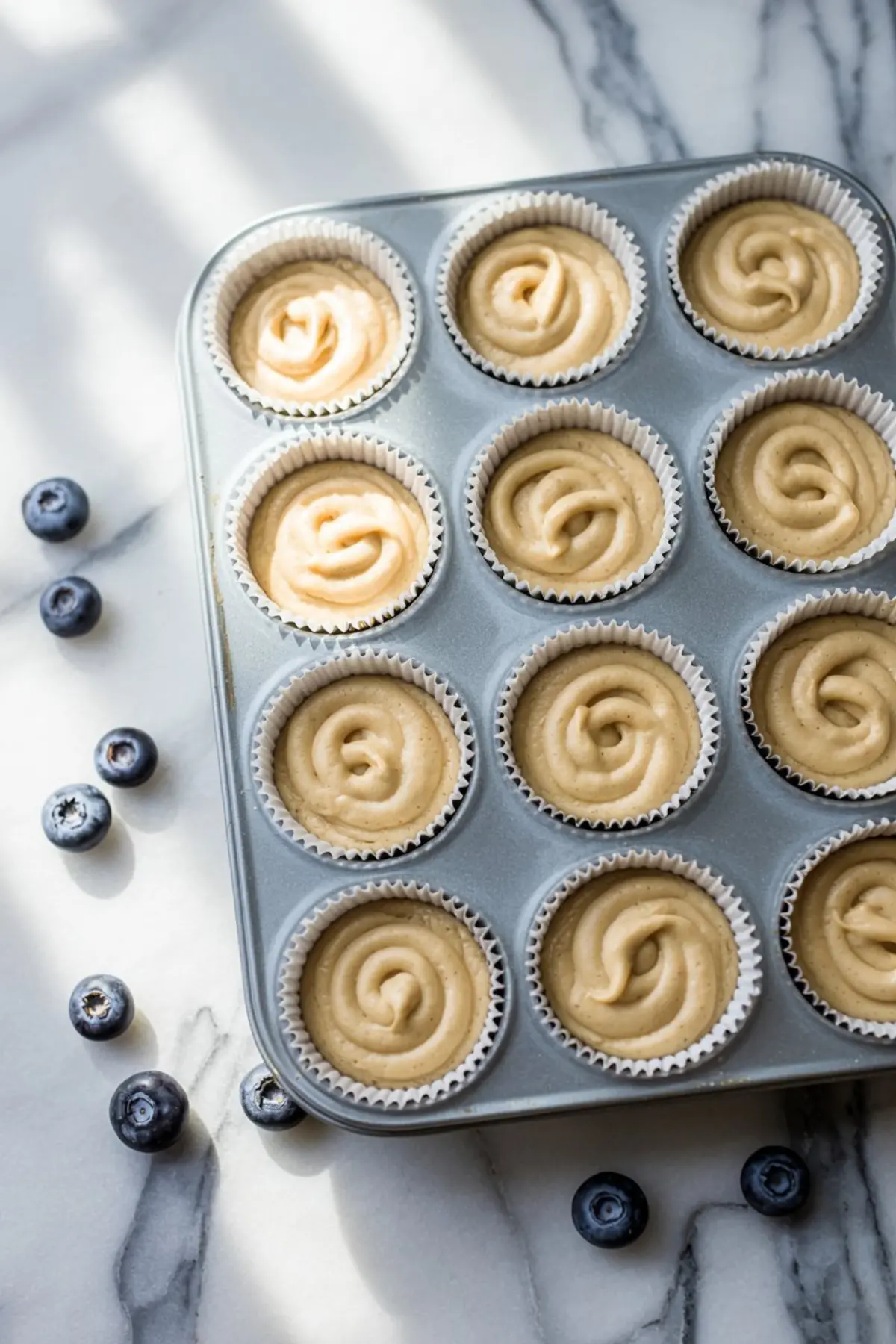 Raw cupcake batter swirled into paper liners in a metal muffin tray, placed on a marble surface with fresh blueberries scattered around.