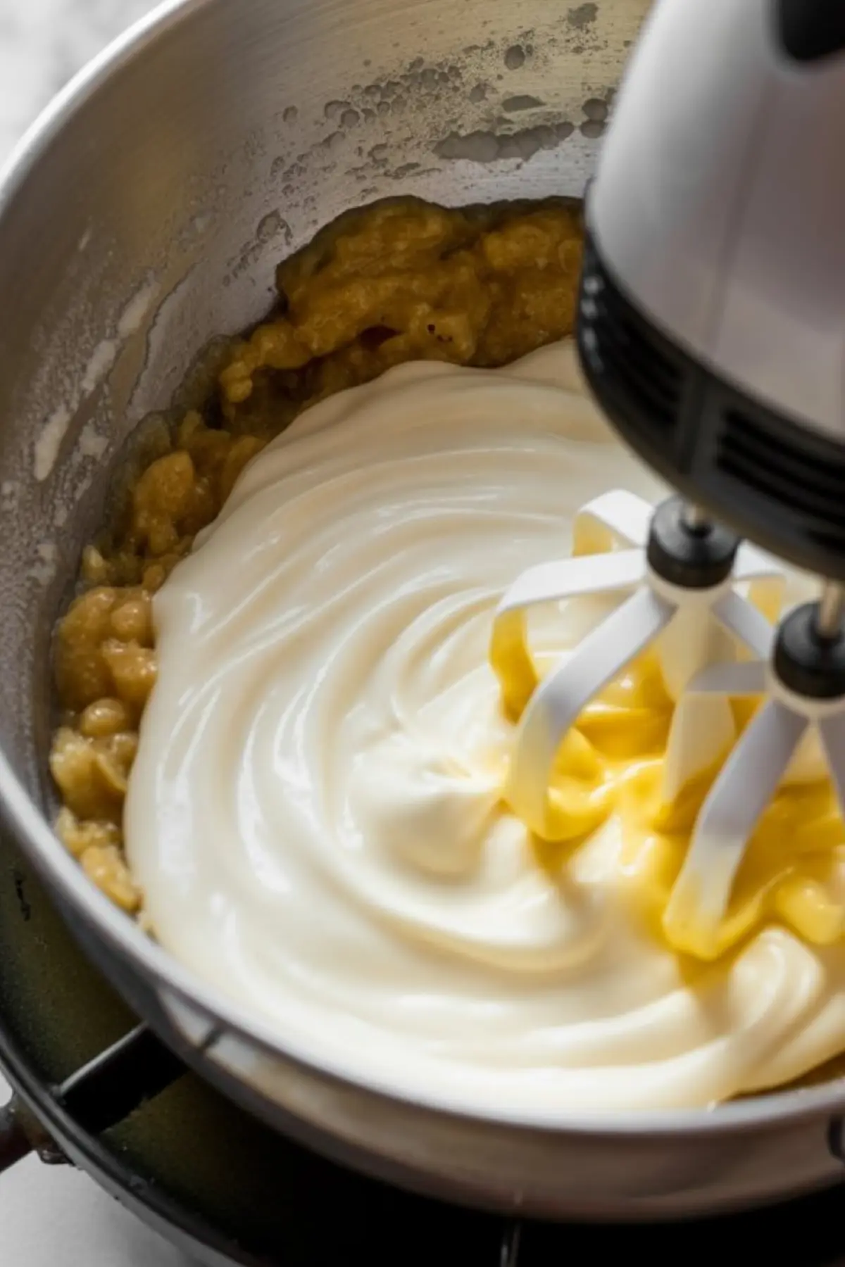 A hand mixer blends creamy white frosting into mashed banana batter inside a metal mixing bowl, showing a close-up of the smooth swirl forming during mixing.
