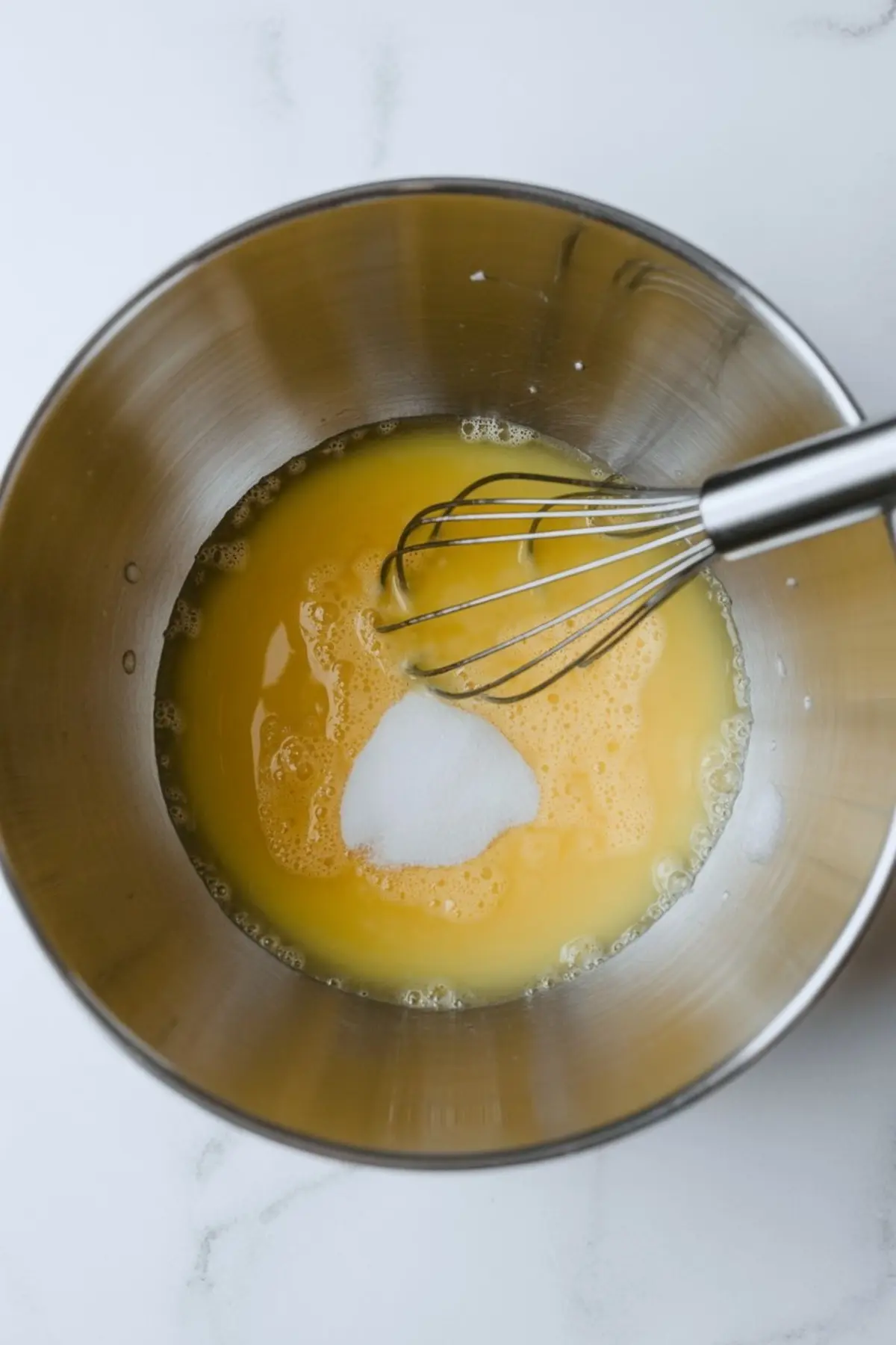Whisk resting in a metal bowl with a mixture of beaten eggs and sugar, ready for blending into a cake or frosting recipe.