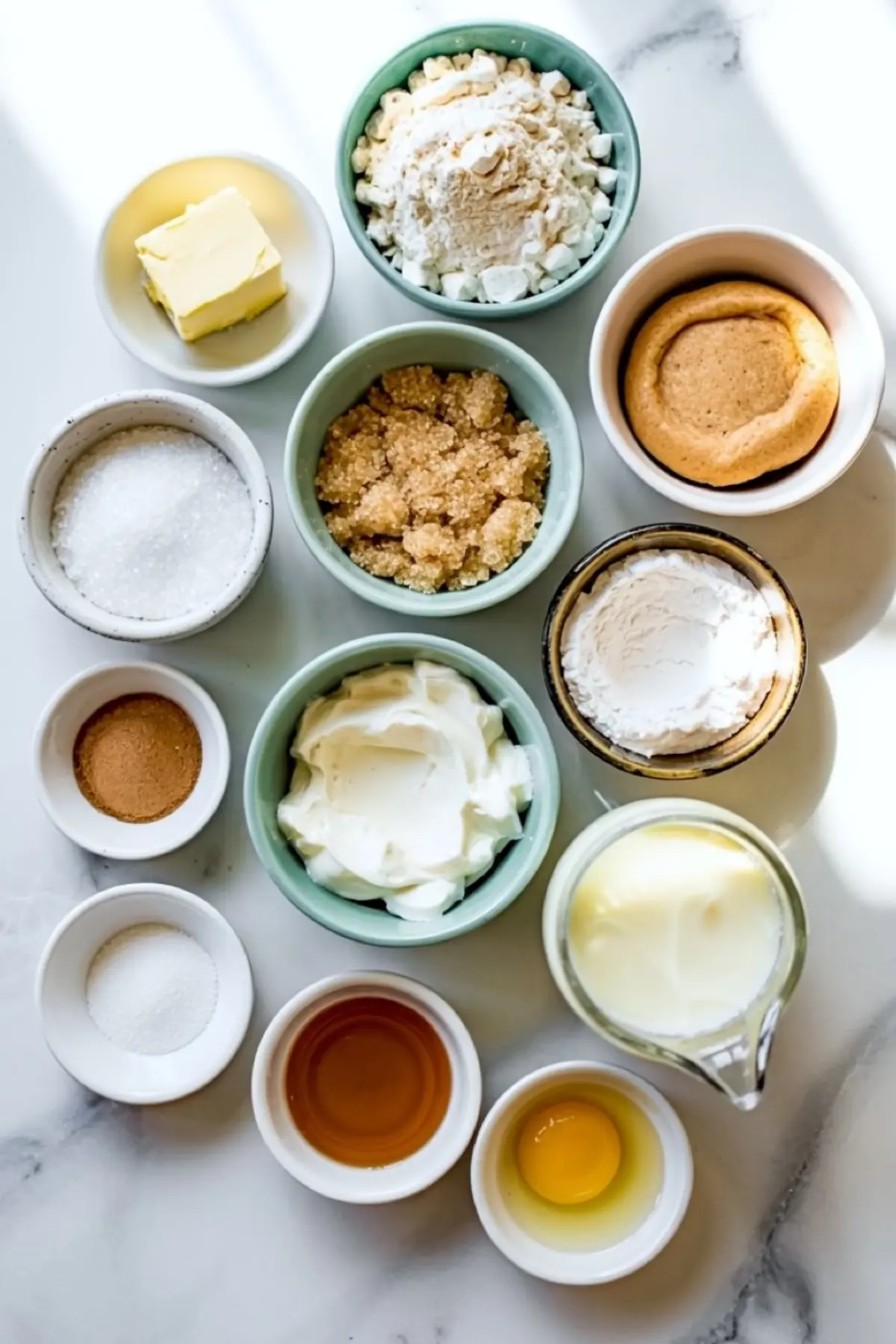 Flat lay of snickerdoodle cupcake ingredients in small bowls, including butter, brown sugar, sour cream, flour, vanilla extract, egg, and cinnamon.