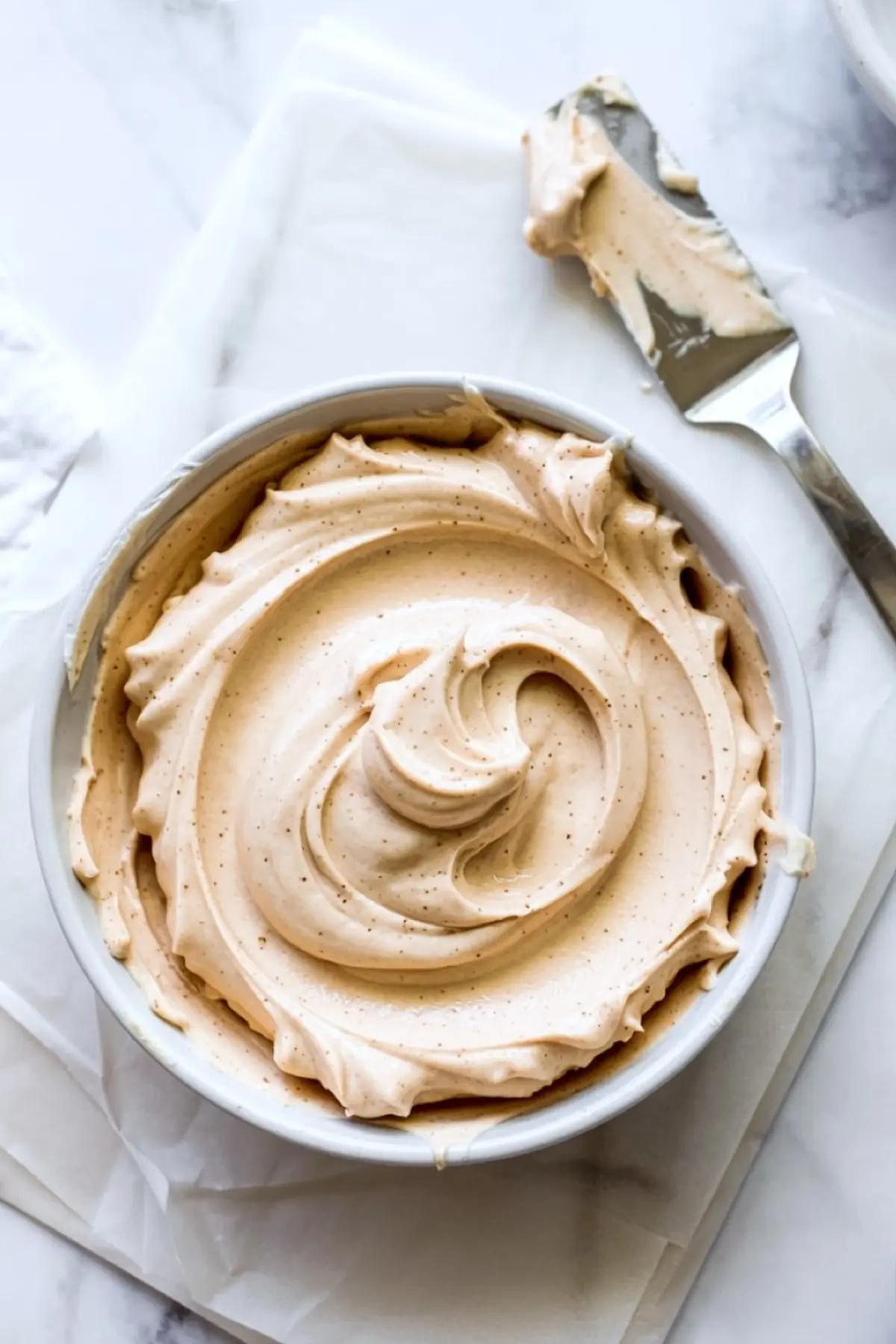 Creamy cinnamon-speckled frosting in a mixing bowl with a metal spatula, ready to be piped onto cupcakes for a sweet snickerdoodle flavor.