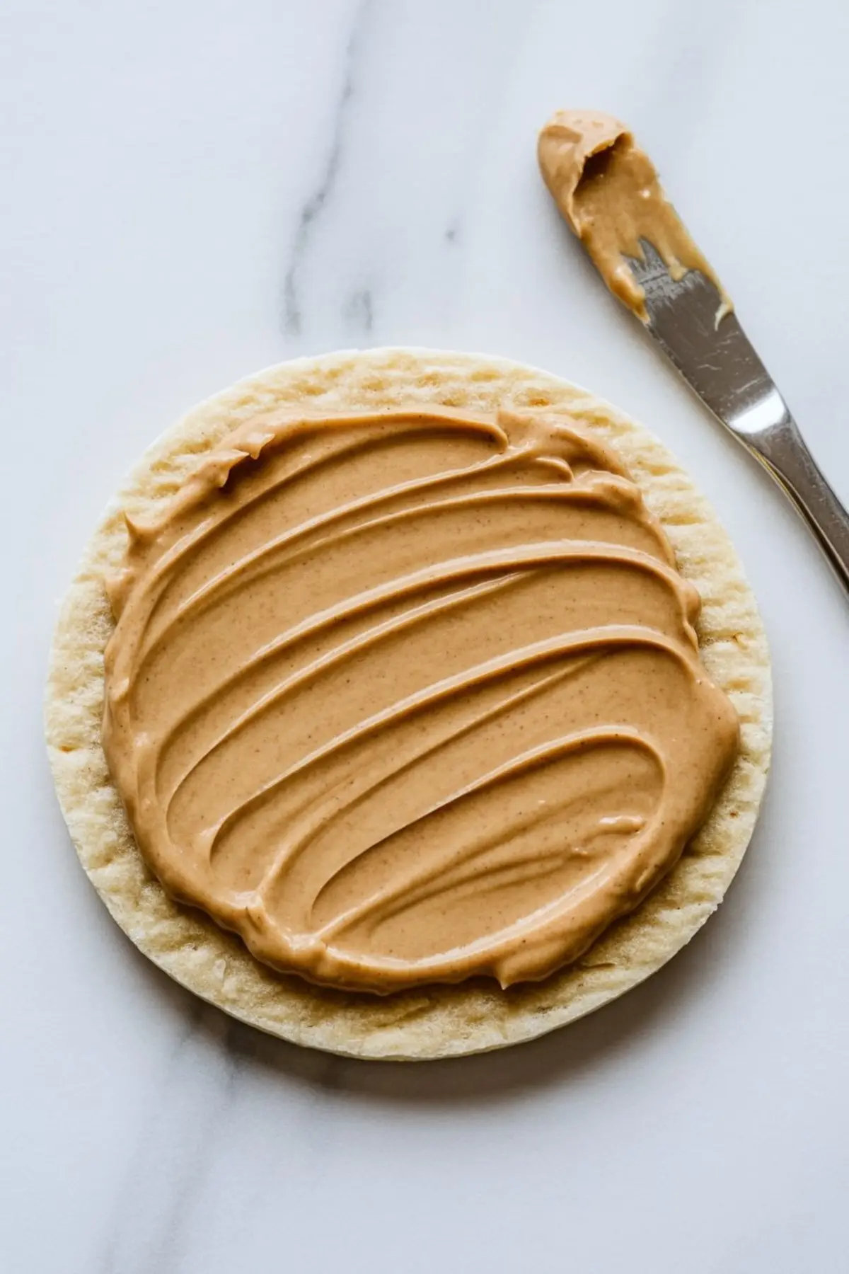 Flatbread round evenly spread with creamy almond butter, placed next to a butter knife on a white marble surface.
