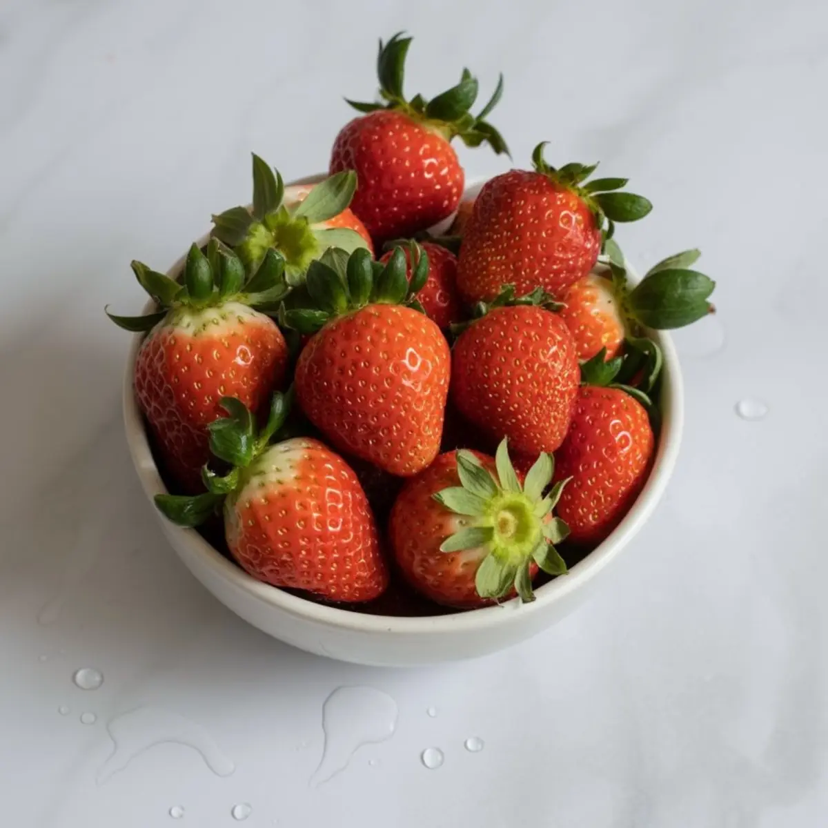 White ceramic bowl packed with fresh, ripe strawberries with green tops on a marble surface with water droplets.
