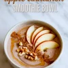 Apple cinnamon smoothie bowl topped with fresh apple slices, ground cinnamon, chopped walnuts, and maple syrup drizzle in a white bowl.