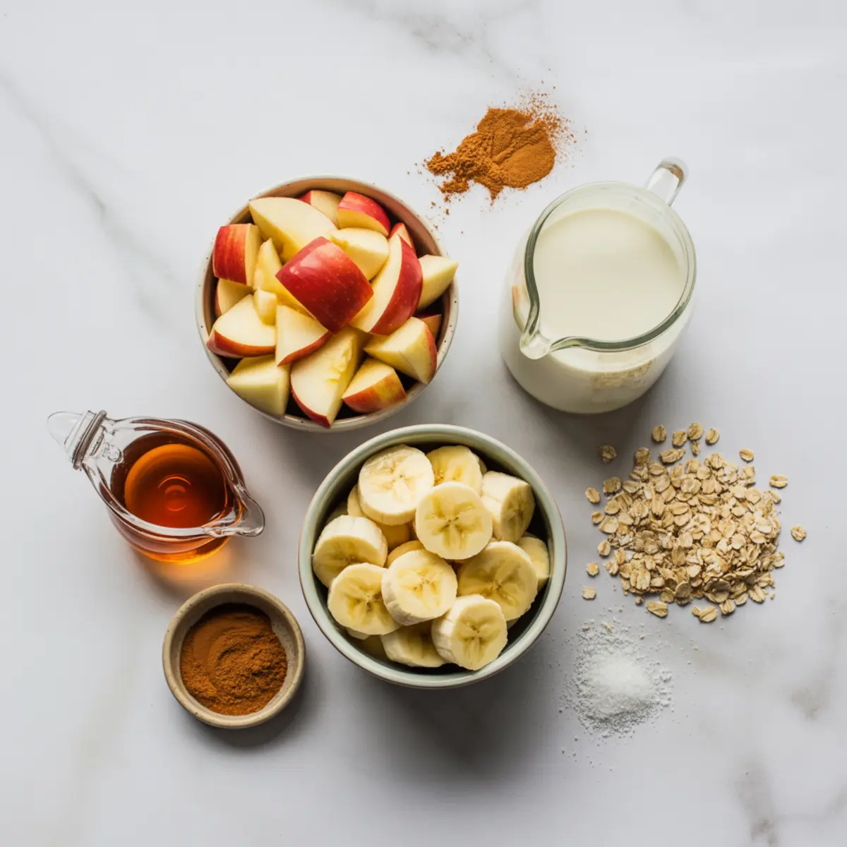 Flat lay of apple cinnamon smoothie ingredients including chopped apples, banana slices, rolled oats, cinnamon, maple syrup, milk, and baking powder on a marble surface.
