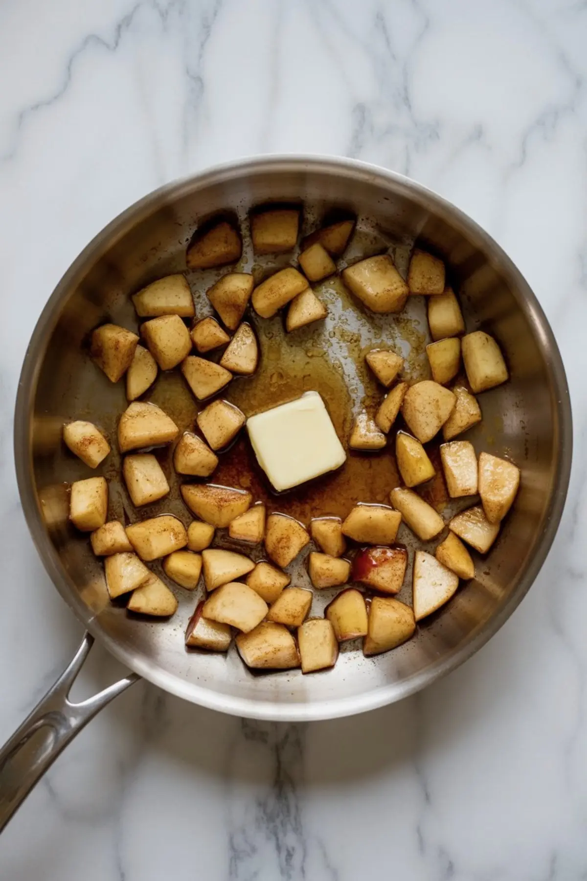 Chopped apples sautéed with butter, cinnamon, and brown sugar in a stainless steel skillet, creating a caramelized apple filling.
