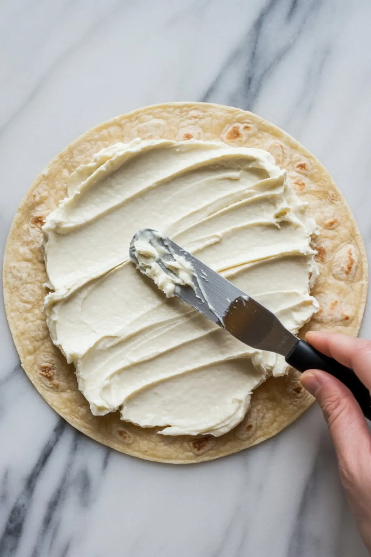 Hand spreading cream cheese mixture evenly over a flour tortilla using an offset spatula on a marble background.
