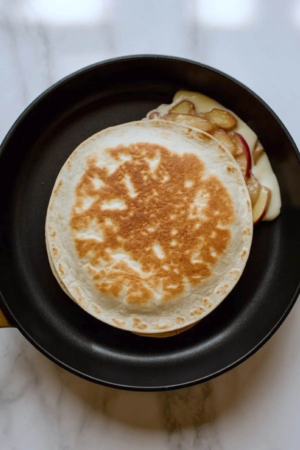 Golden brown apple quesadilla in a skillet with caramelized apple filling and melted cream cheese slightly oozing out from the side.
