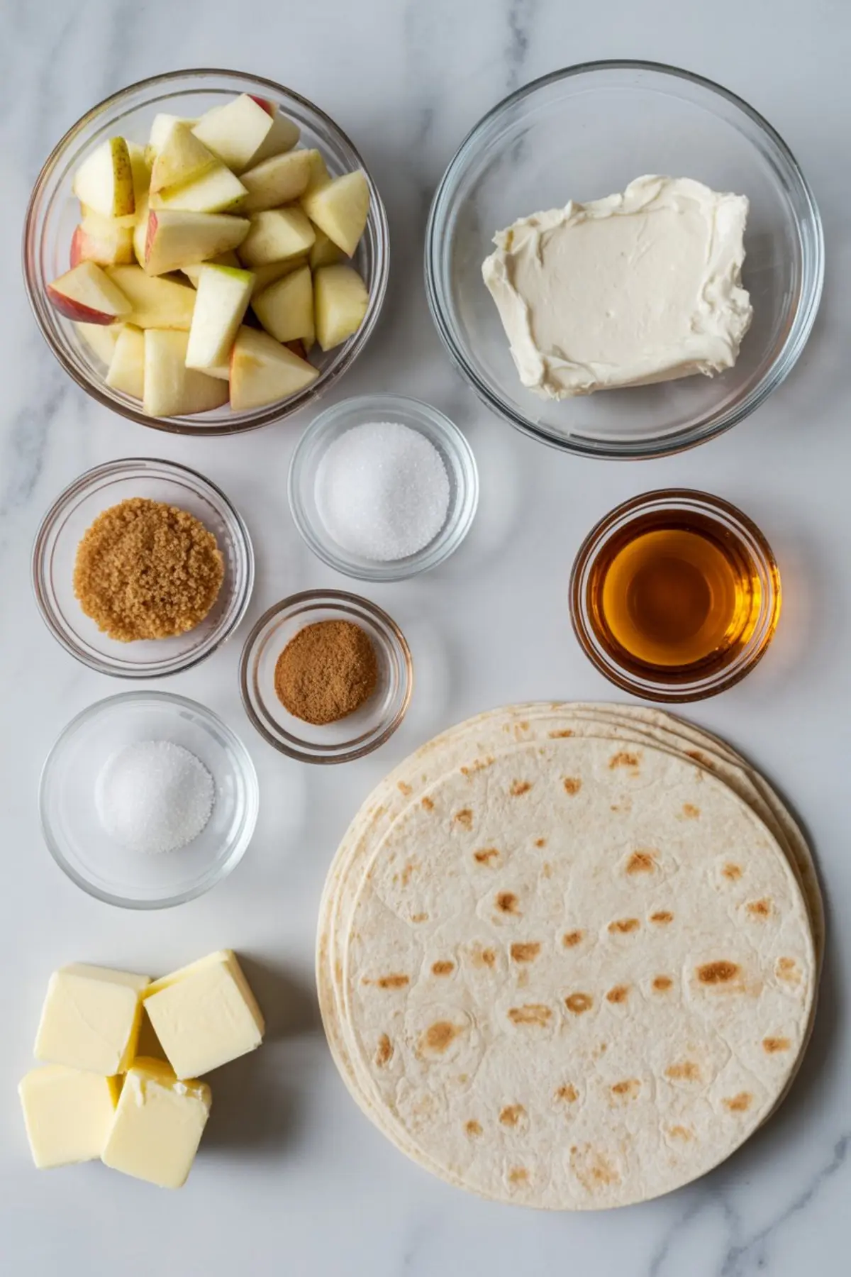 Overhead view of recipe ingredients on a marble surface including chopped apples, cream cheese, butter cubes, brown sugar, white sugar, cinnamon, tortillas, and maple syrup arranged in glass bowls.
