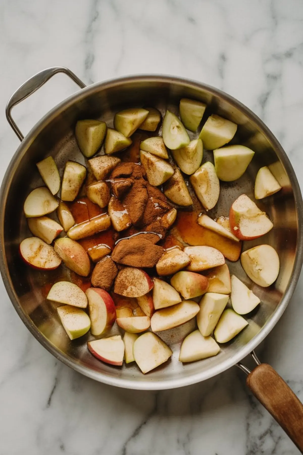 Sliced apples in a stainless steel skillet with cinnamon, maple syrup, and spices, prepped for cooking on a marble countertop.
