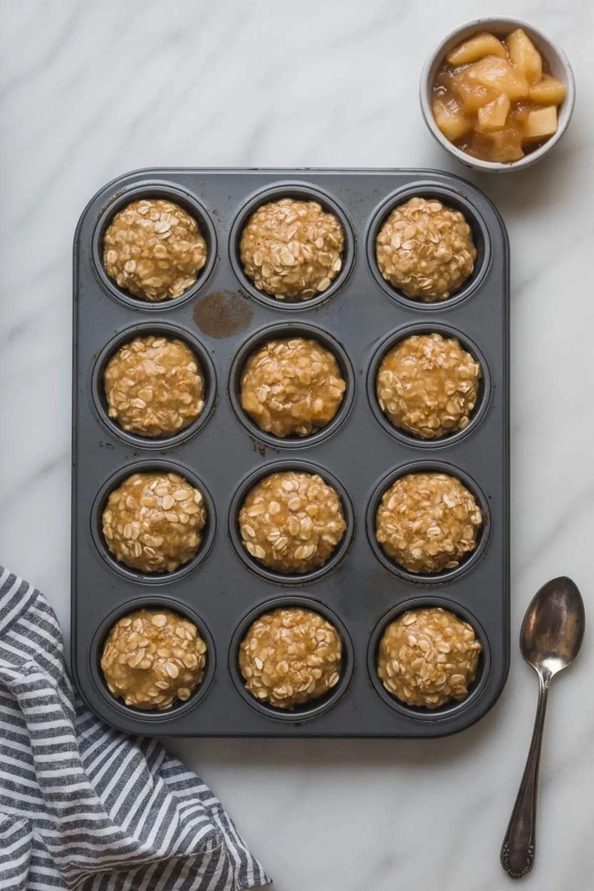 Unbaked apple pie oatmeal mixture in a nonstick muffin tin, each cavity filled with oat batter, with a bowl of cooked apple filling and a vintage spoon placed on the side.
