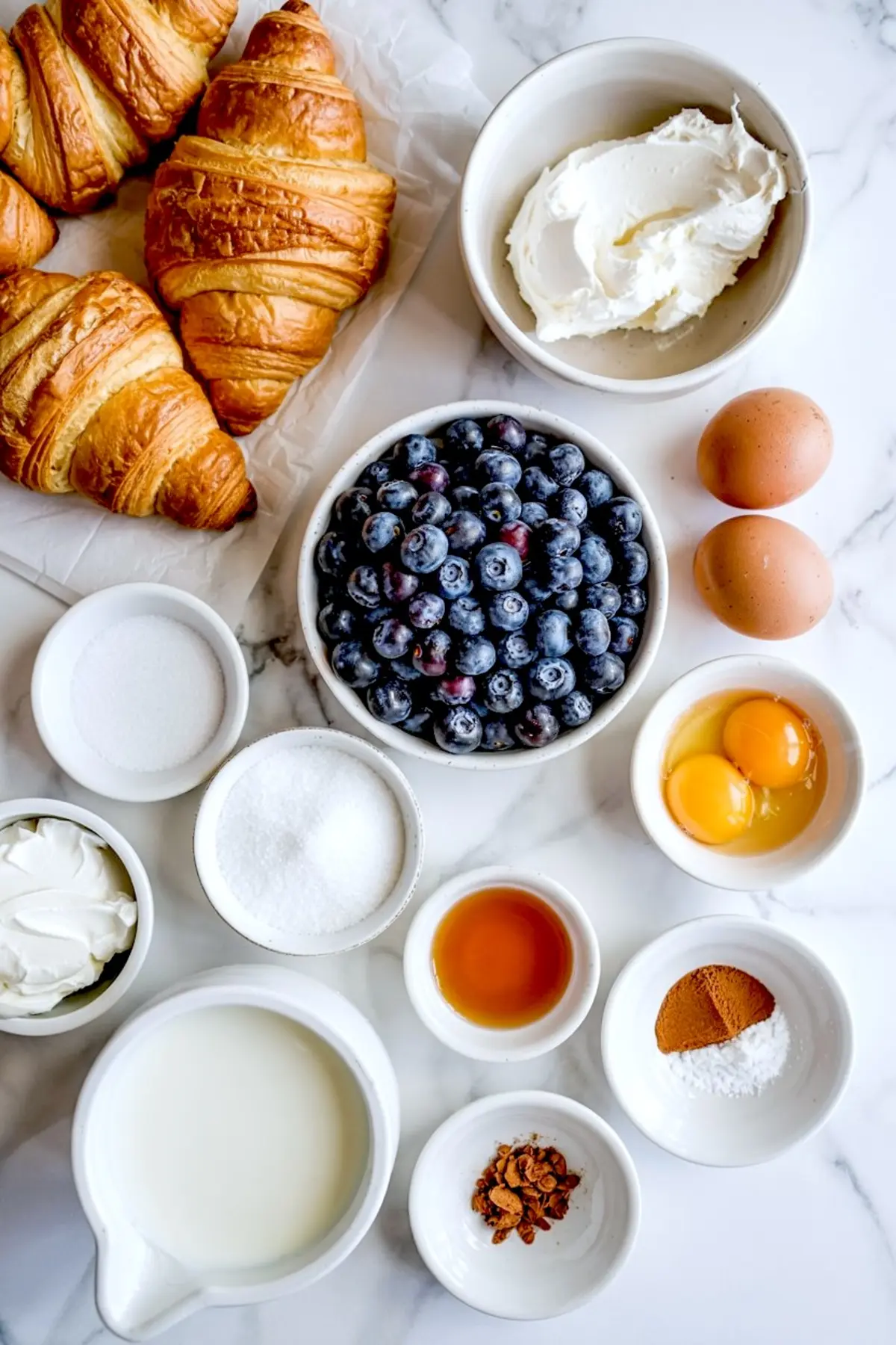 Flat lay of fresh baking ingredients including croissants, blueberries, eggs, cream cheese, milk, sugar, and vanilla extract on a white marble countertop.