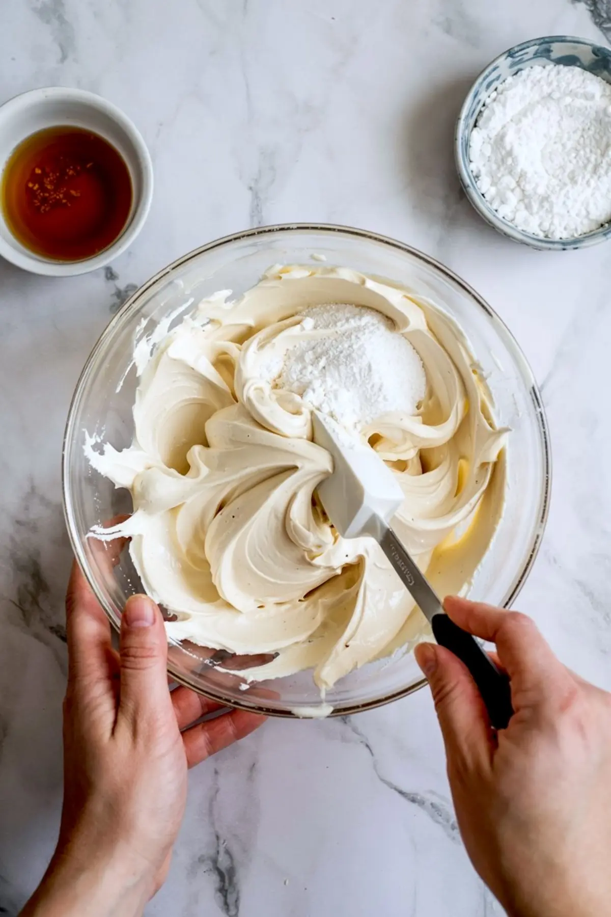 Hands mixing powdered sugar into a creamy vanilla batter using a white spatula in a glass bowl, with bowls of vanilla extract and powdered sugar on a marble countertop.